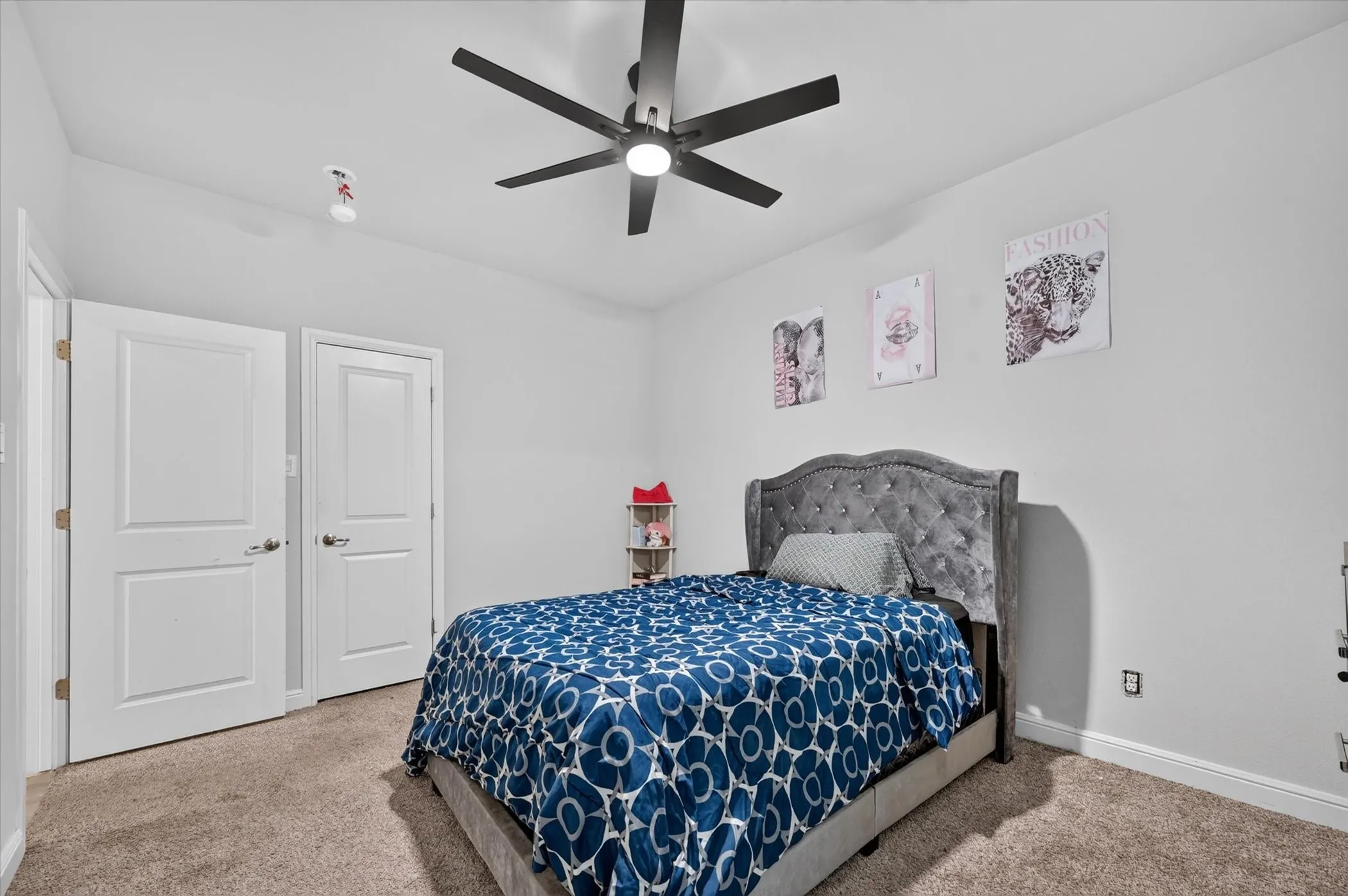 Spacious bedroom featuring a tufted upholstered headboard, plush carpeting, ceiling fan with integrated lighting, dual closets with six-panel doors, and soft neutral wall finishes accented with botanical artwork