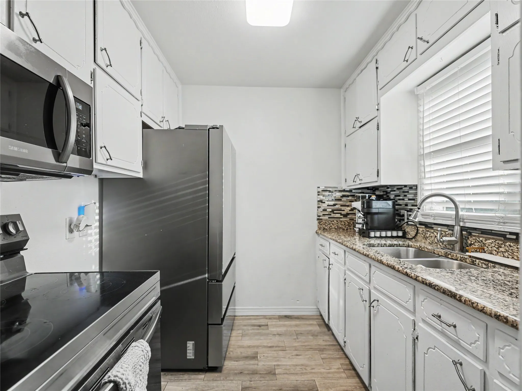 Galley kitchen with white cabinetry, wood-finish flooring, stainless steel appliances, a multi-tone tile backsplash, and a double basin sink