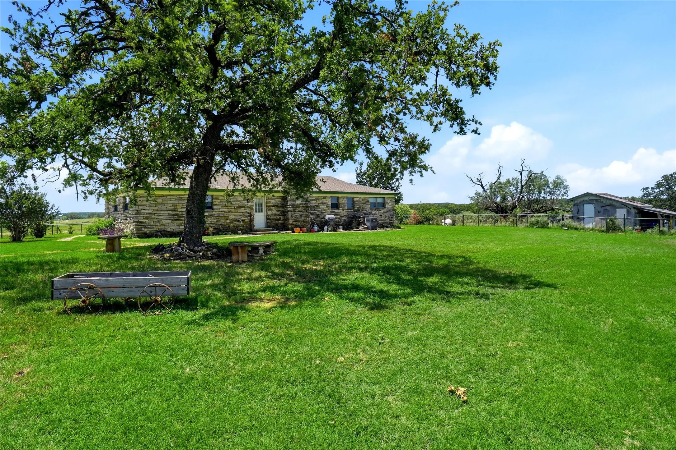 Expansive green lawn with mature trees, a stone-clad single-story residence, and an auxiliary structure