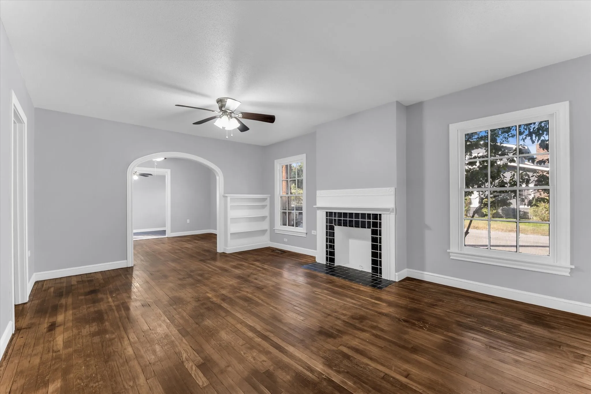 Hardwood flooring throughout, a tiled fireplace with white mantel, built-in shelving, an arched doorway, and a ceiling fan