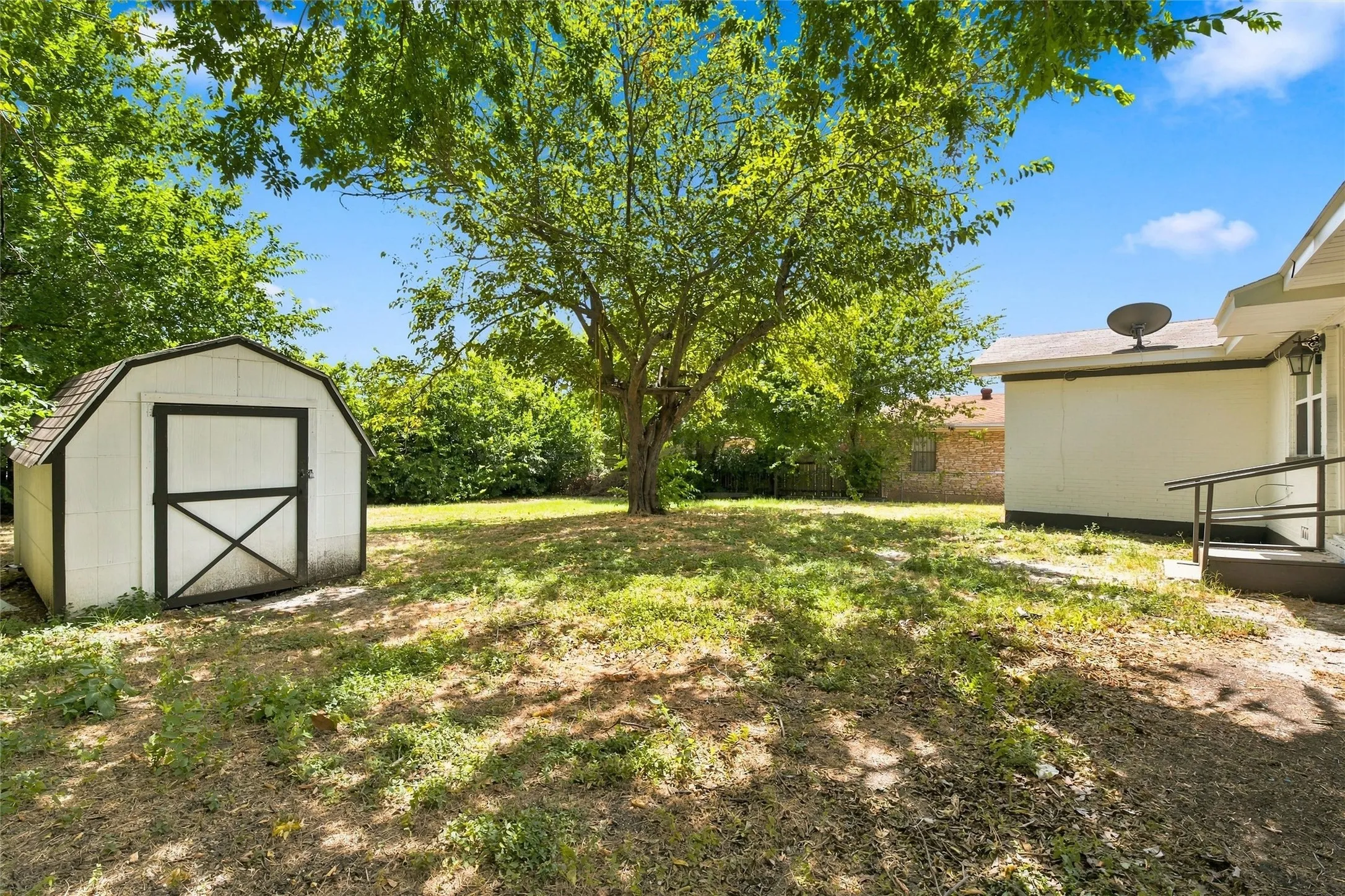 Expansive backyard featuring a white storage shed with dark trim, mature trees, and a ground-level concrete patio with a metal railing
