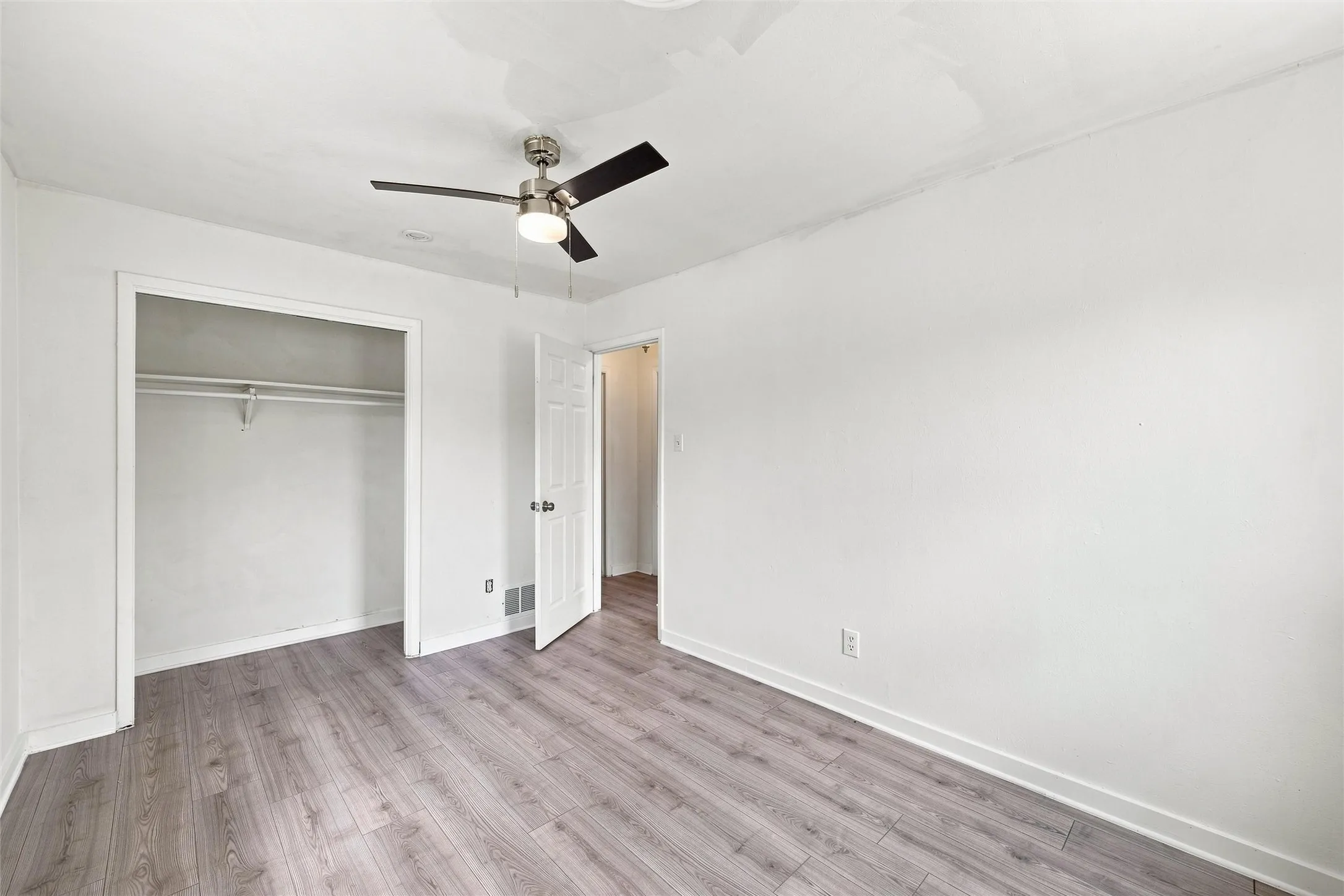 Gray wood-finish flooring, white painted walls, ceiling fan with integrated lighting, and a closet with shelving