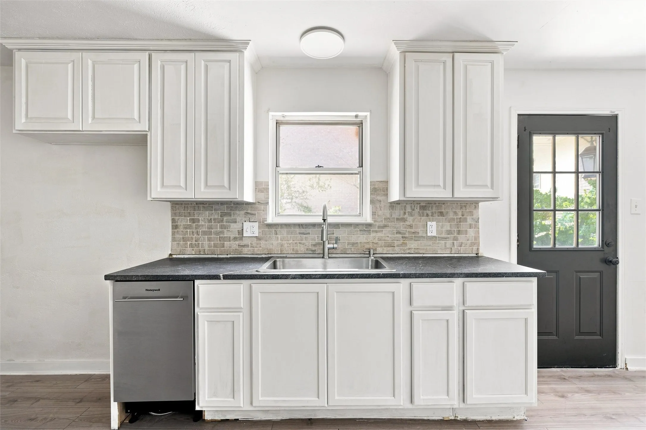 White shaker-style cabinetry with crown molding, stainless steel sink with dual basins, mosaic tile backsplash, and dark countertops