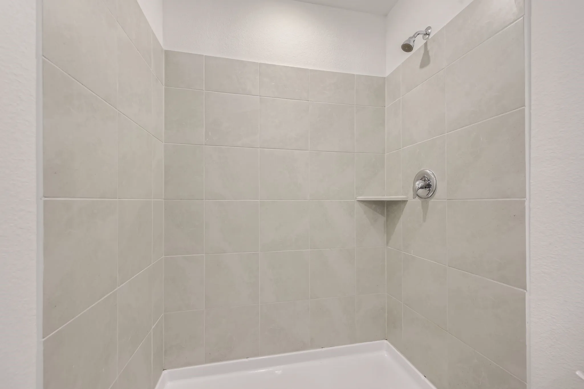 Tiled shower enclosure featuring a built-in shelf, chrome fixtures, and a white shower pan
