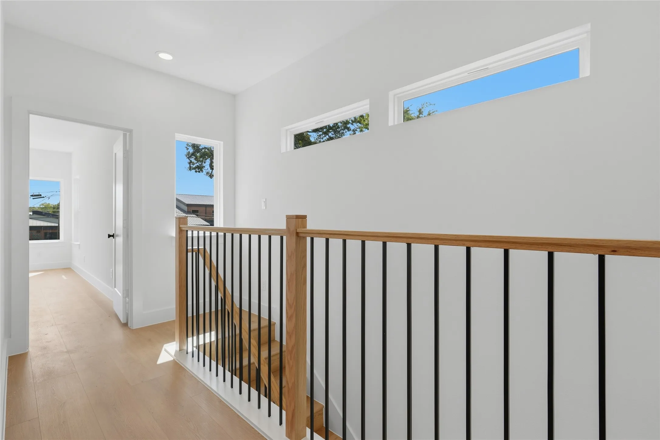 Light wood-finish flooring, contemporary black metal and wood balustrade, recessed ceiling lighting, multiple windows providing natural illumination, and white interior walls