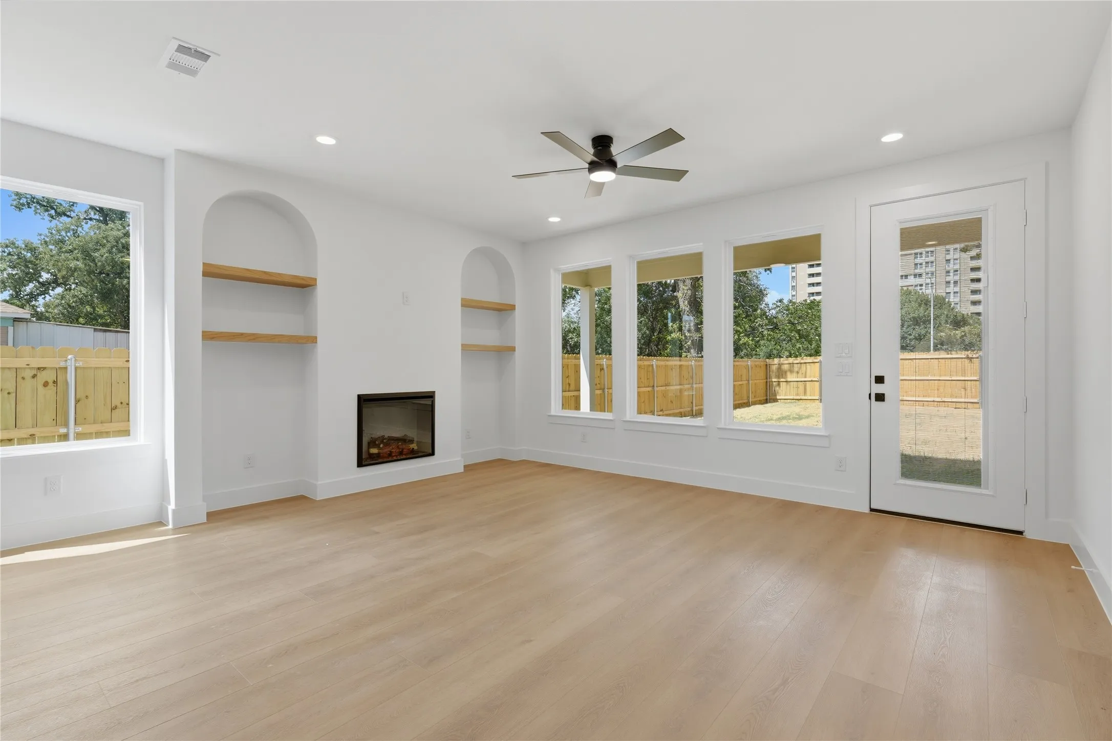 Spacious living area featuring wood-finish flooring, an electric fireplace, two arched built-in shelving units, a ceiling fan with integrated lighting, and multiple windows and a glass-paneled door opening to the exterior