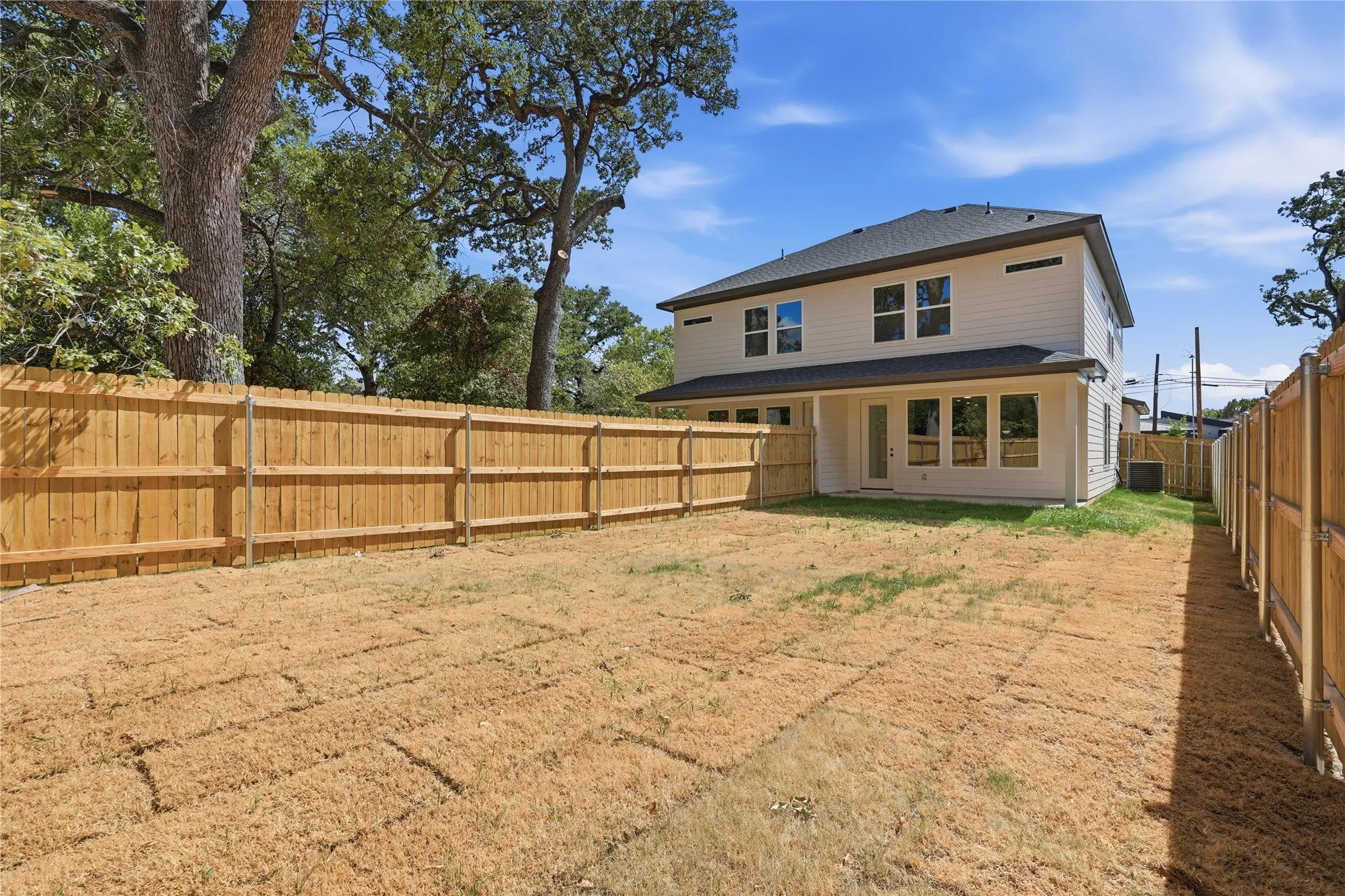 Expansive backyard with a full privacy fence, established trees, and a two-story residence featuring multiple windows and a rear entry door