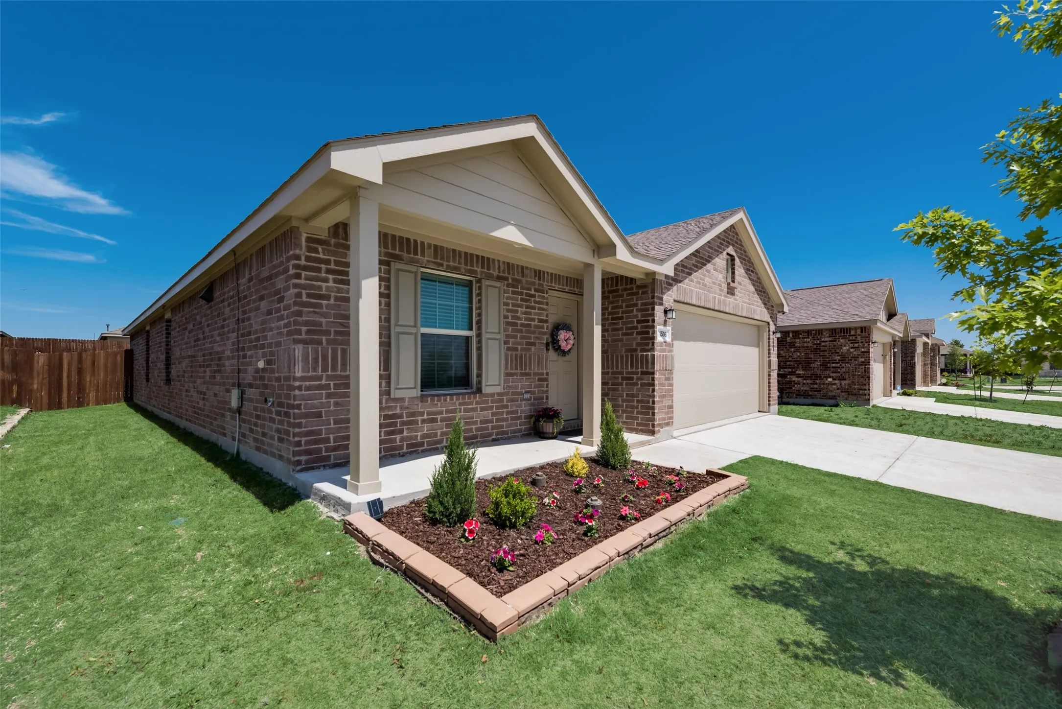 Charming brick residence featuring a covered front porch with white columns, manicured landscaping with mulch beds and ornamental plantings, attached two-car garage, and expansive level lawn with mature shade trees