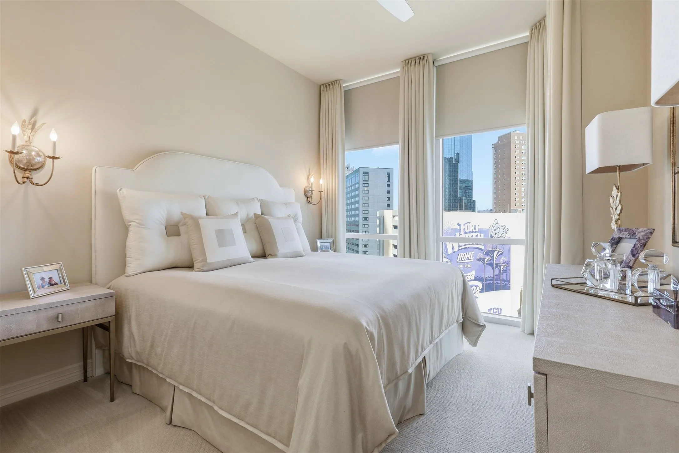 Upholstered headboard with integrated sconces, dual windows with roller shades and full-length drapes, and a ceiling fan