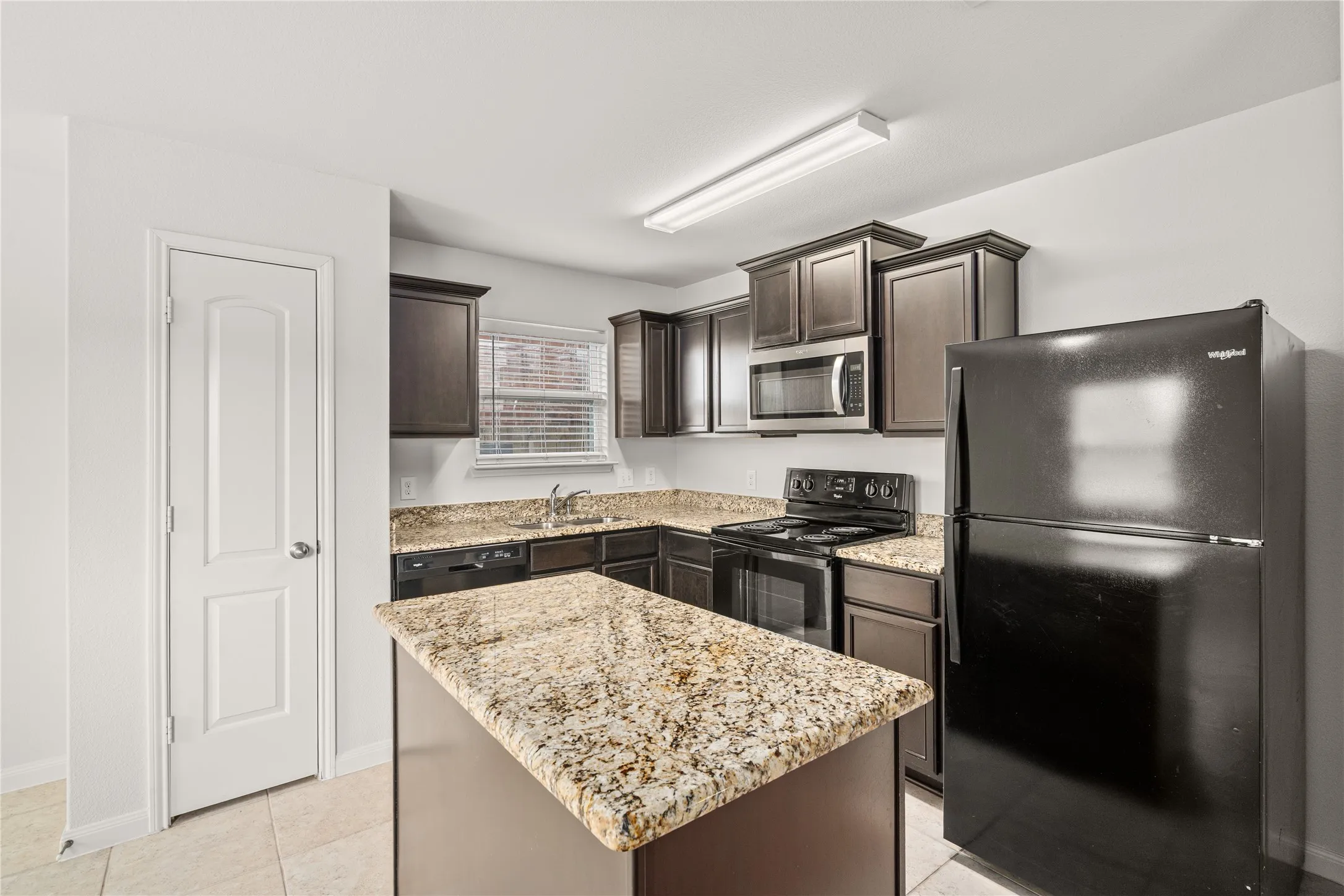 Kitchen featuring an island, black appliances, dark wood-finish cabinetry, granite countertops, and tile flooring