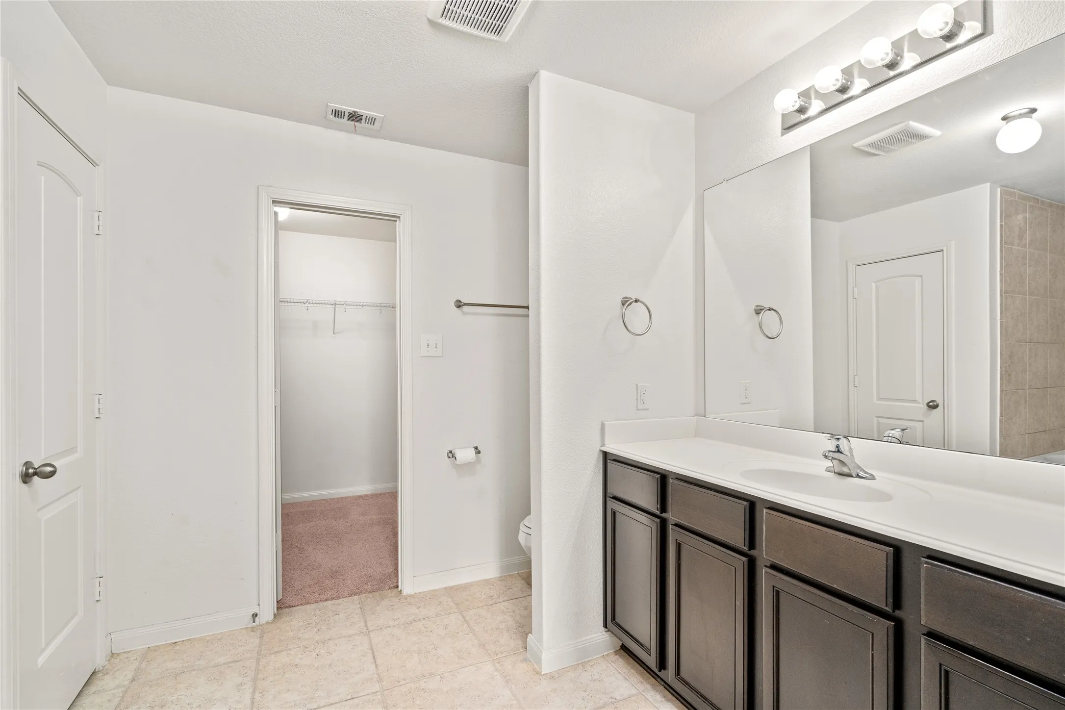 Tile flooring throughout, ample vanity with dual sinks, overhead vanity lighting, and a walk-in closet with carpeted flooring