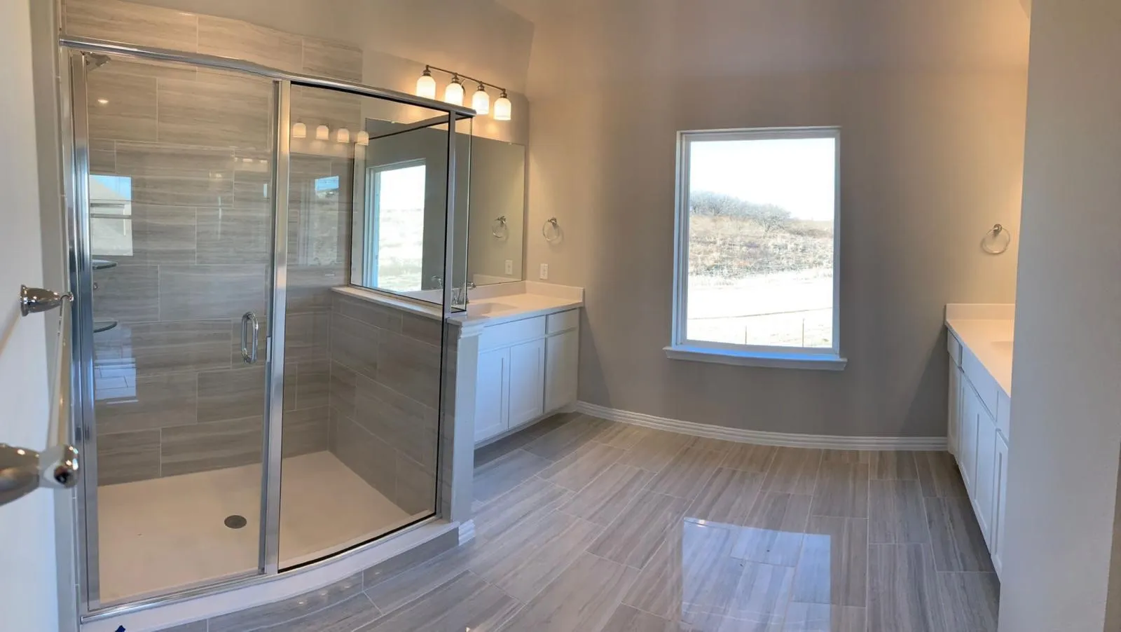 Glass-enclosed shower with tiled walls and a white base, dual vanities with white cabinetry and light countertops, and wood-finish tile flooring