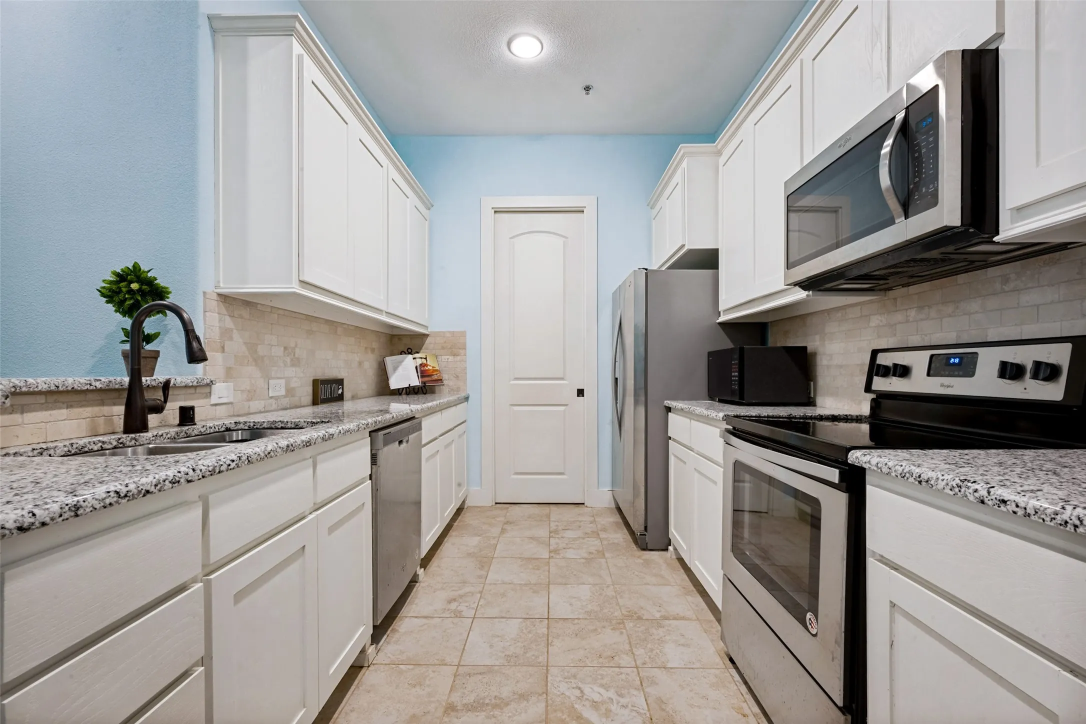 Gallery kitchen featuring white cabinetry, light-colored granite counter tops, stainless steel appliances, a tile backsplash, and tile flooring