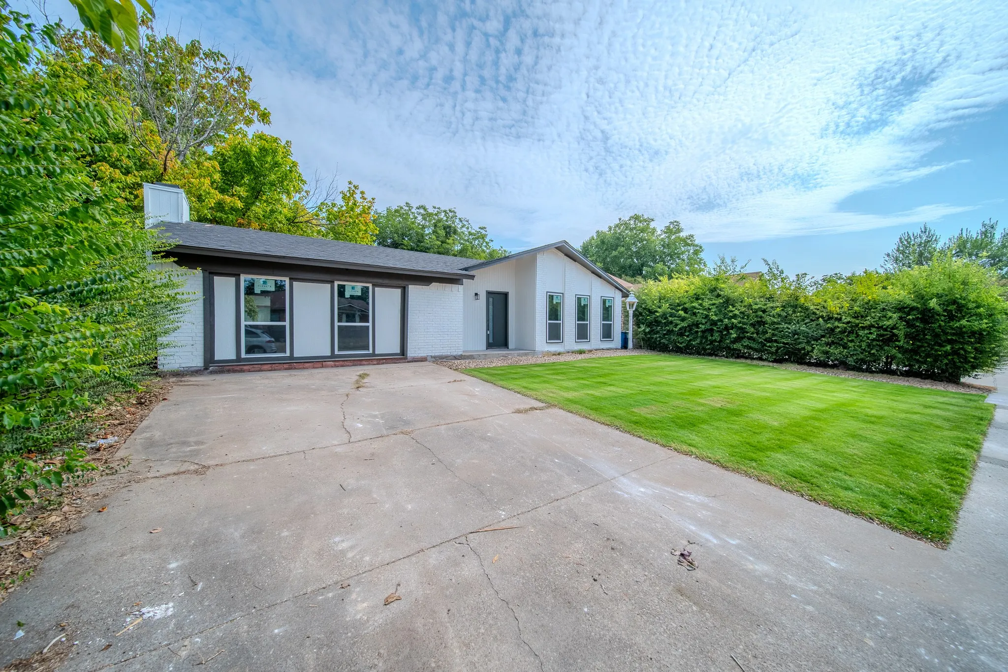 White brick exterior, dark trim windows, dark shingle roof, established green landscaping, concrete driveway