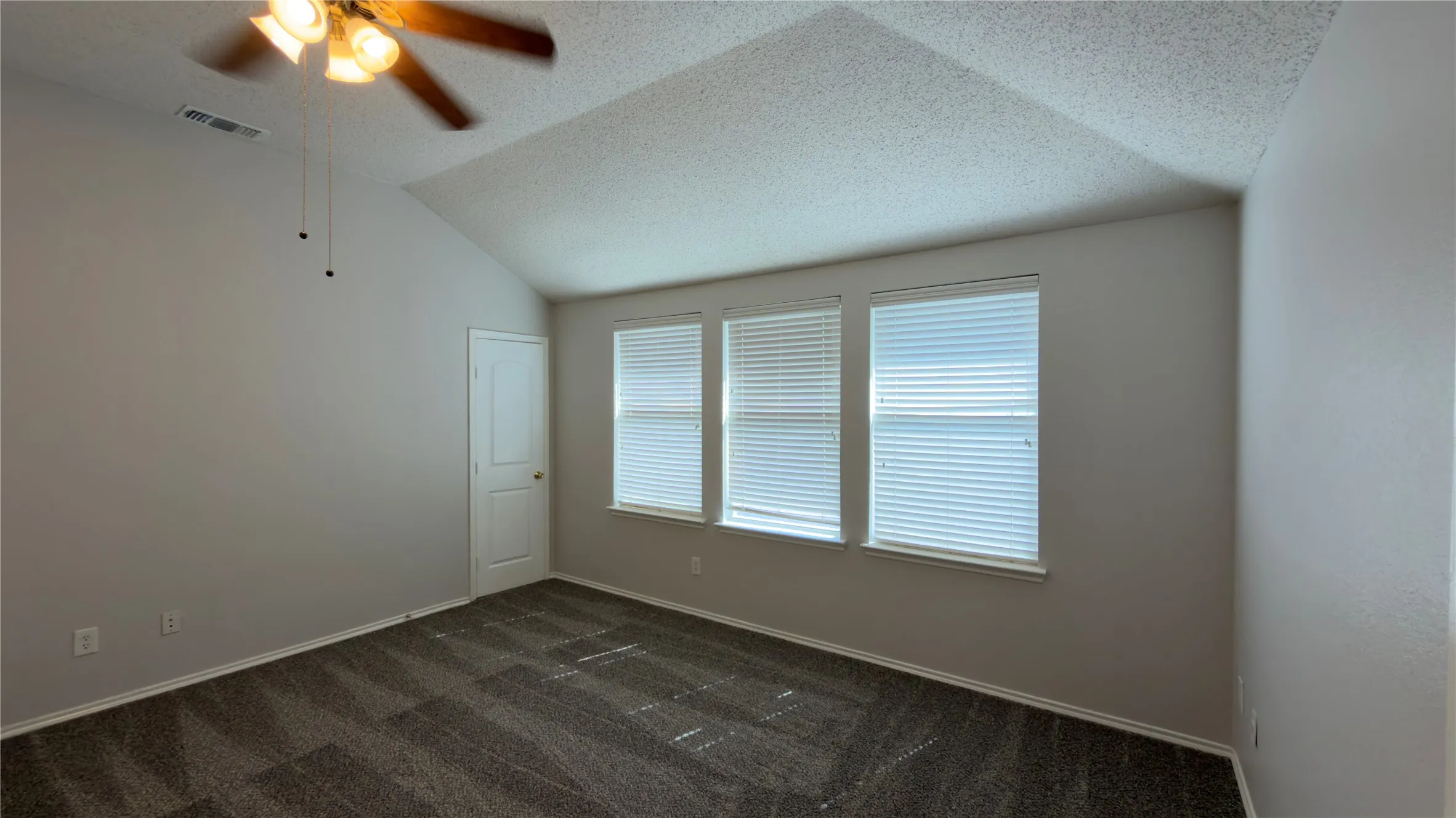 Vaulted ceiling, ceiling fan with light fixture, three windows with blinds, interior door, and carpet flooring