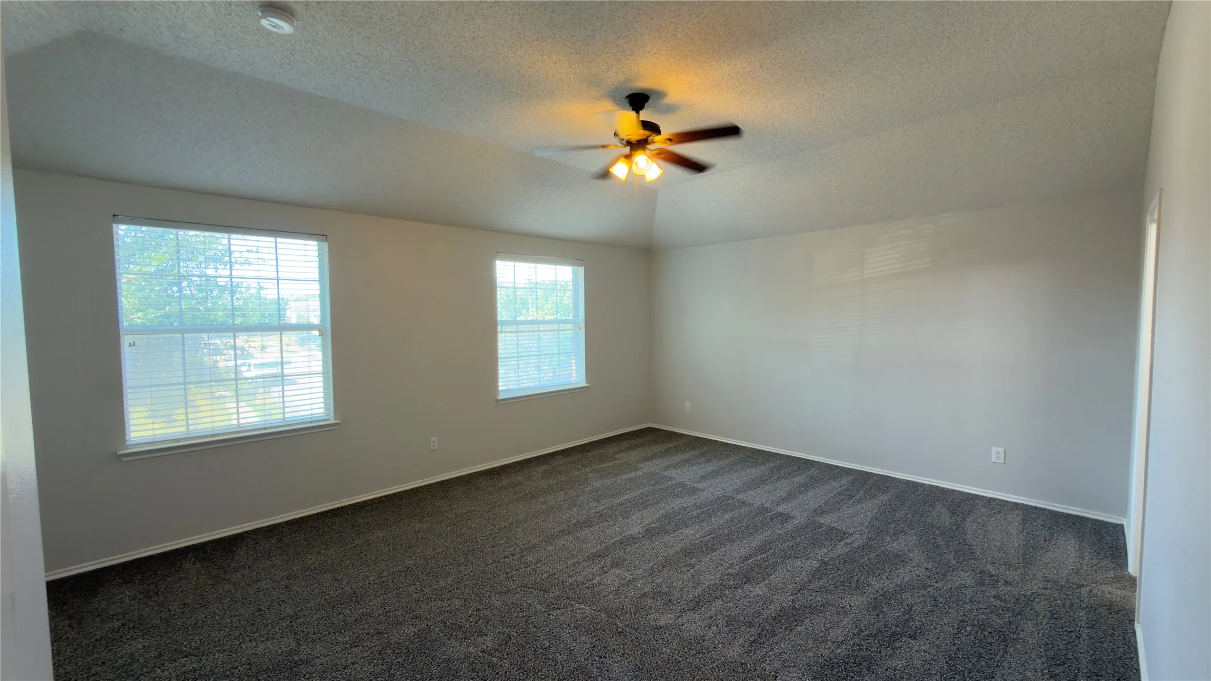 Vaulted ceiling with an attached ceiling fan, dual windows, and textured carpet flooring