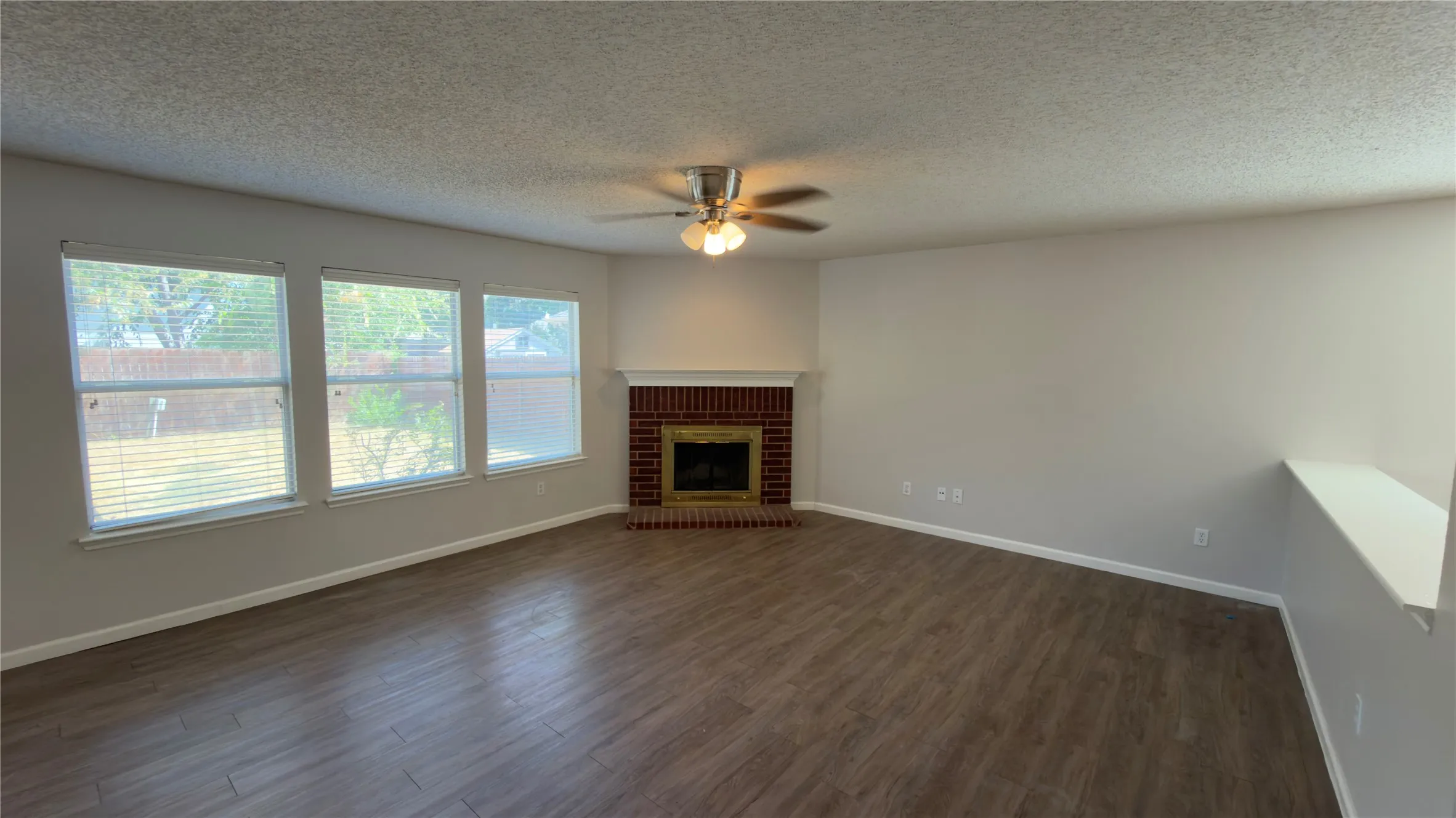 Wood-finish flooring throughout, multiple windows, corner fireplace with brick surround, and a ceiling fan with integrated lighting