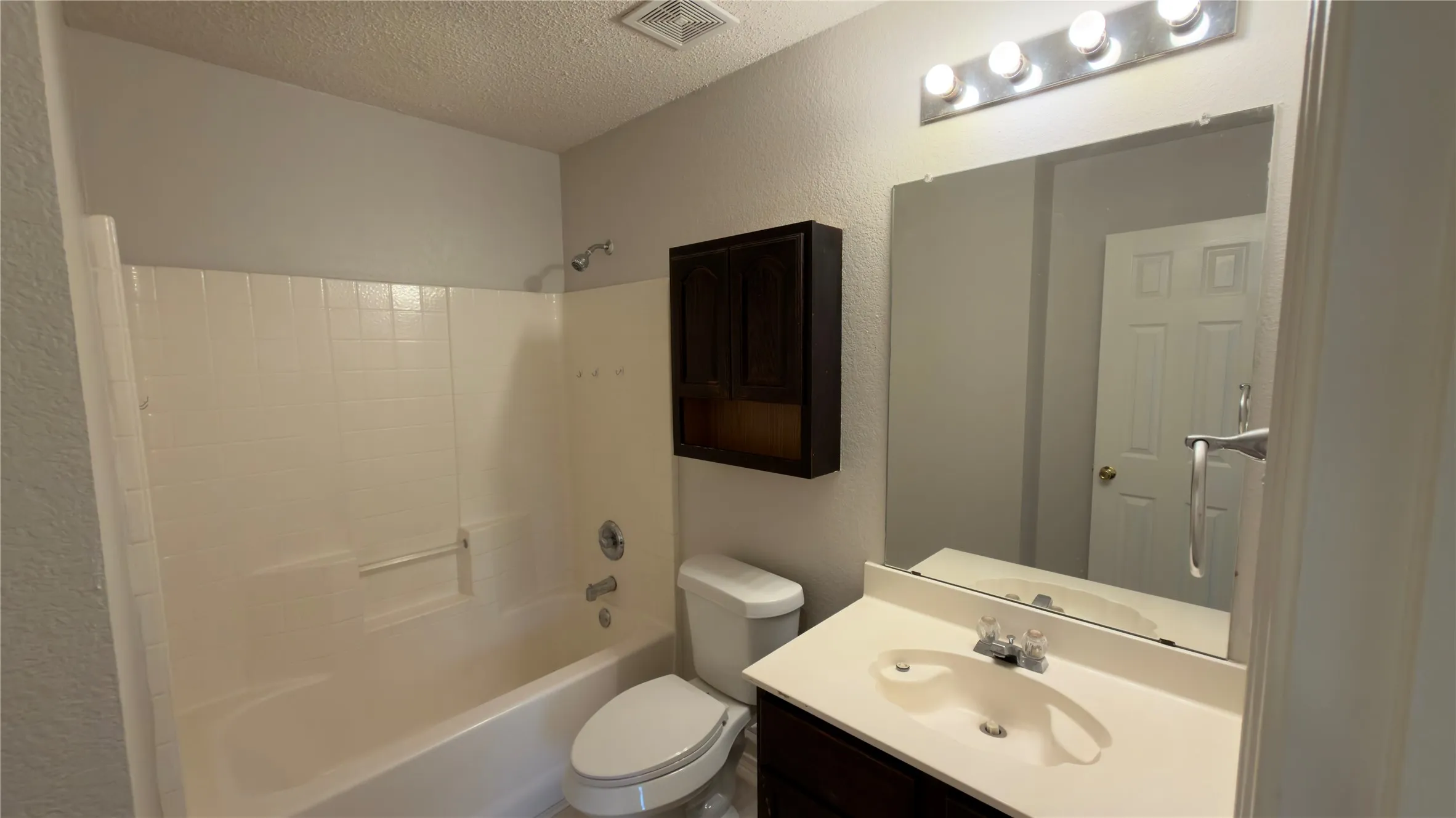 White bathtub with integrated shower, dark wood-finish vanity with a white countertop, chrome-finish fixtures, and a large wall-mounted mirror