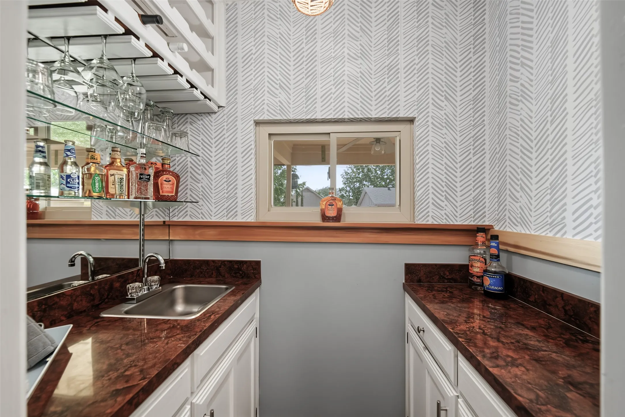 Built-in wet bar featuring a stainless steel sink, brown countertop, white cabinetry, and overhead glass storage
