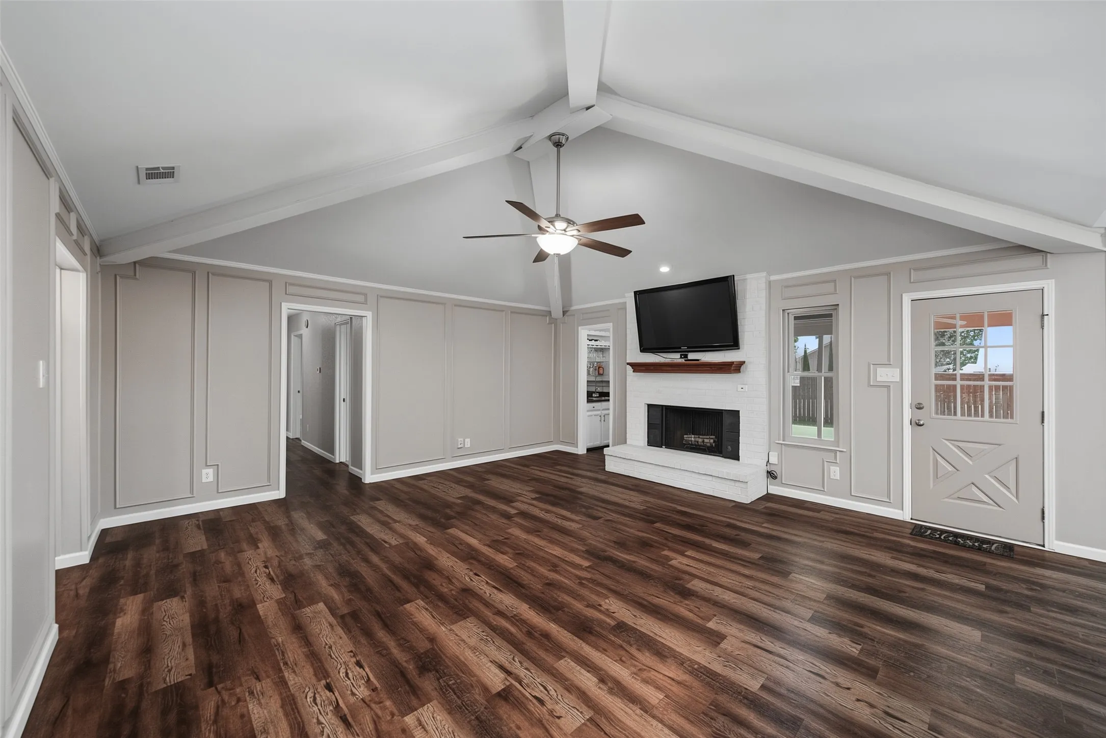 Vaulted ceiling with exposed beams, wood-finish flooring, white brick fireplace with wood mantel, ceiling fan, and decorative wall paneling