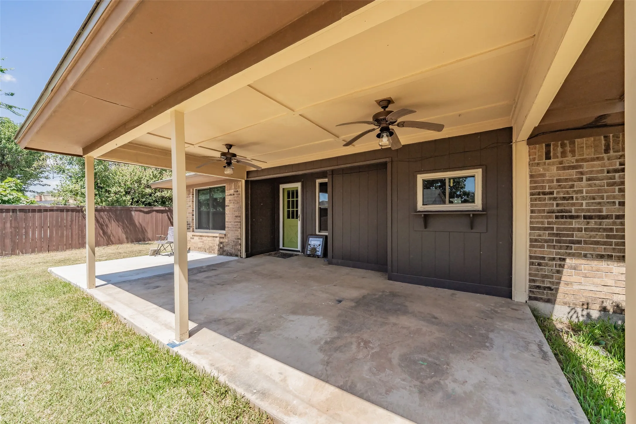 Expansive covered patio featuring two ceiling fans with integrated lighting, a concrete slab, a green paneled door, and a brown vertical siding exterior