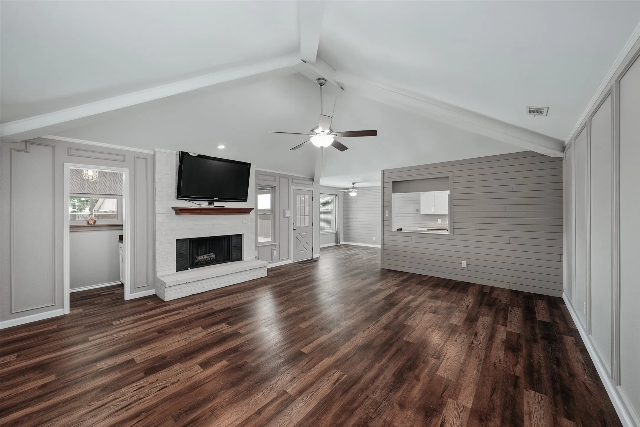 Vaulted ceiling with exposed beams, wood-finish flooring, white brick fireplace with mantel, ceiling fan.