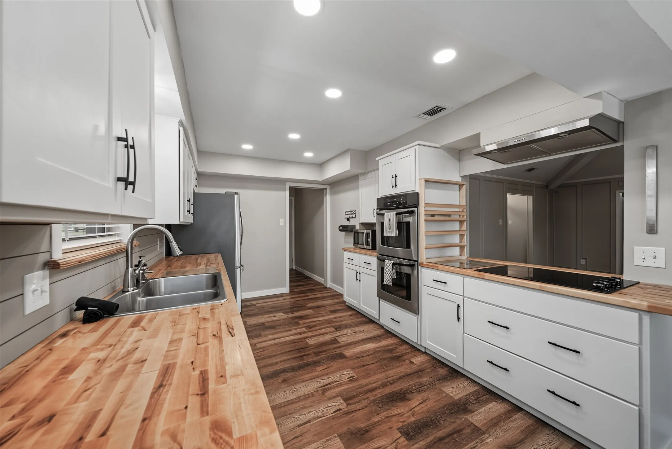 Kitchen featuring luxury vinyl plank flooring, white cabinetry with matte black hardware, integrated double ovens, a stainless steel range hood, and butcher block countertops