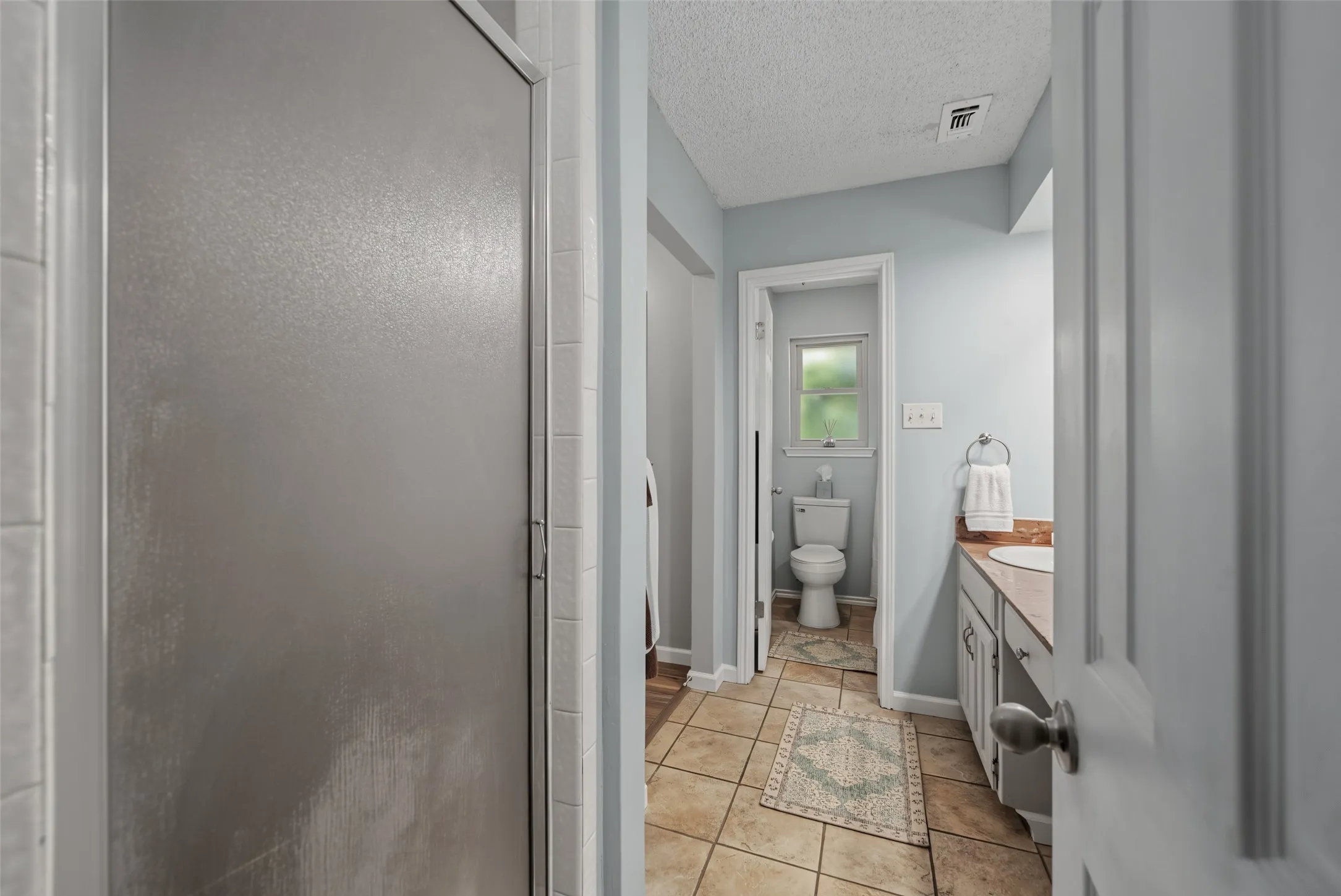 Primary bath: Frosted glass shower enclosure, tiled flooring, vanity with a light-colored countertop, and a toilet with a window above