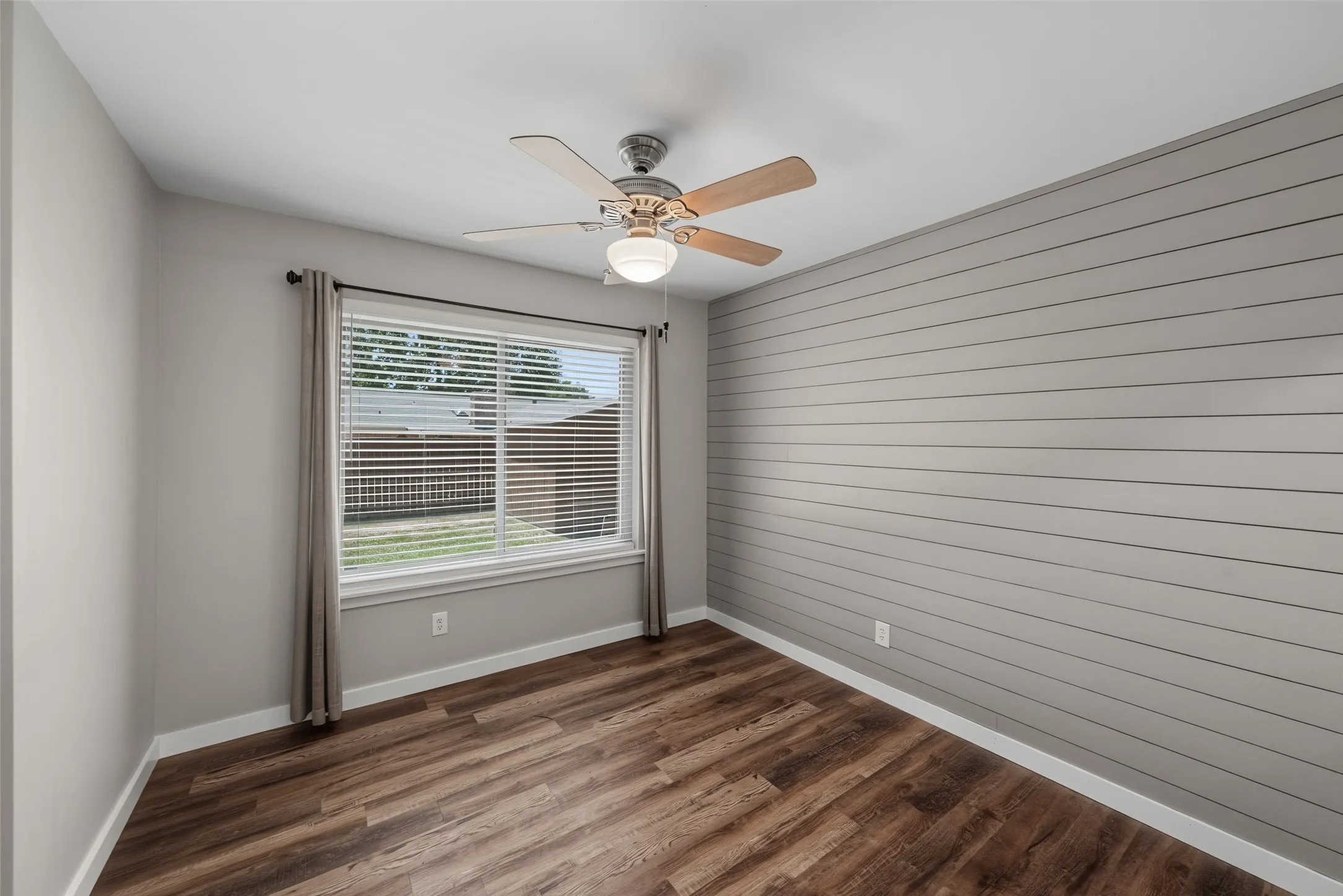 Wood-finish flooring, accent wall with horizontal paneling, ceiling fan with light fixture, and window with blinds