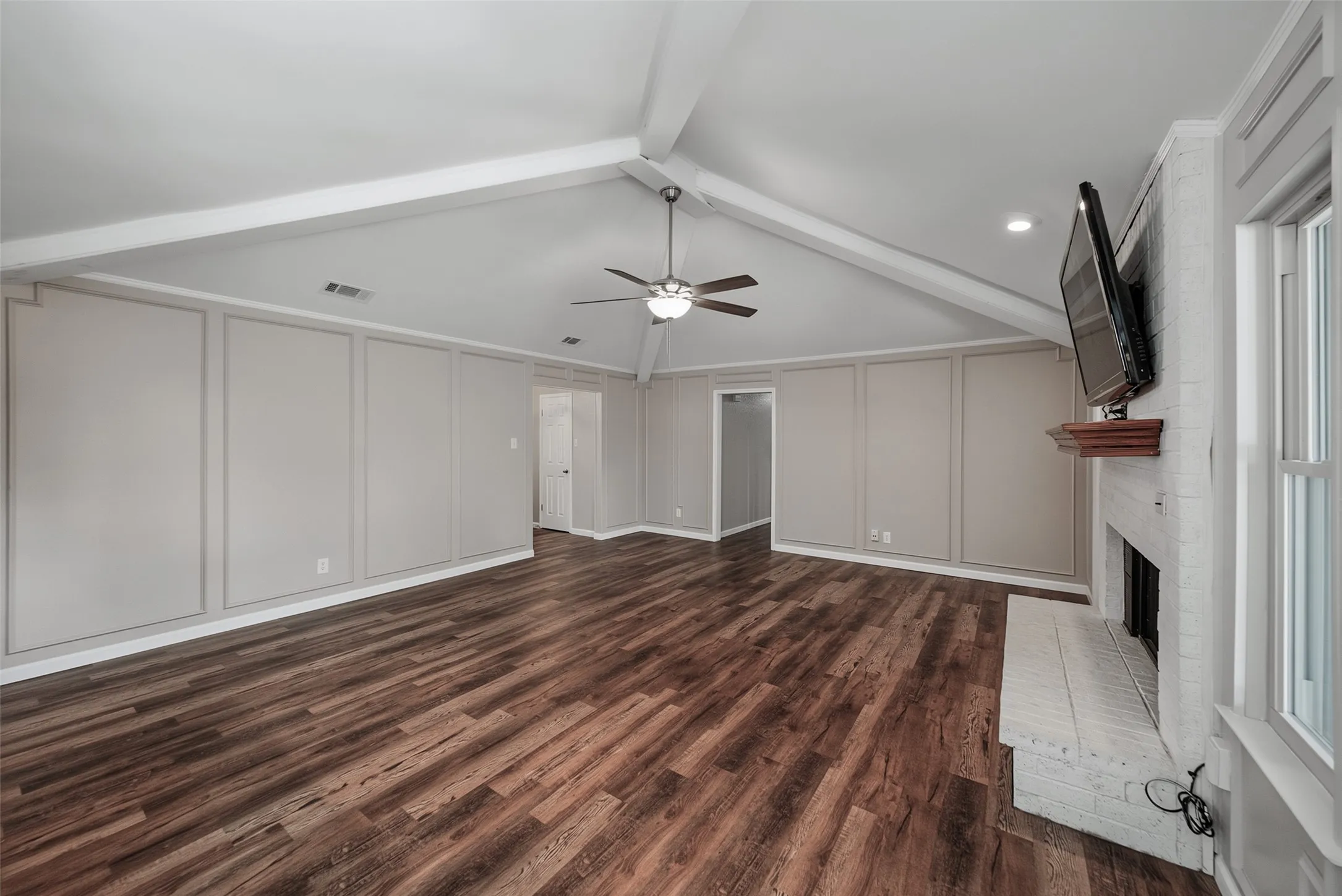 Vaulted ceiling with exposed beams, wood-finish flooring, accent wall paneling, brick fireplace with wood mantel, and ceiling fan
