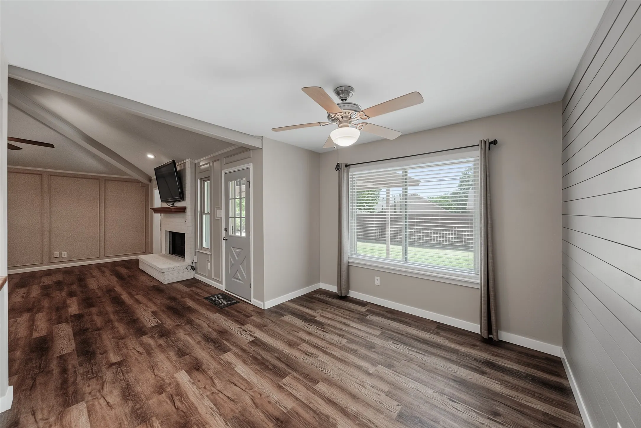 Wood-finish flooring throughout, a ceiling fan with integrated lighting, a spacious window with blinds, and a built-in fireplace