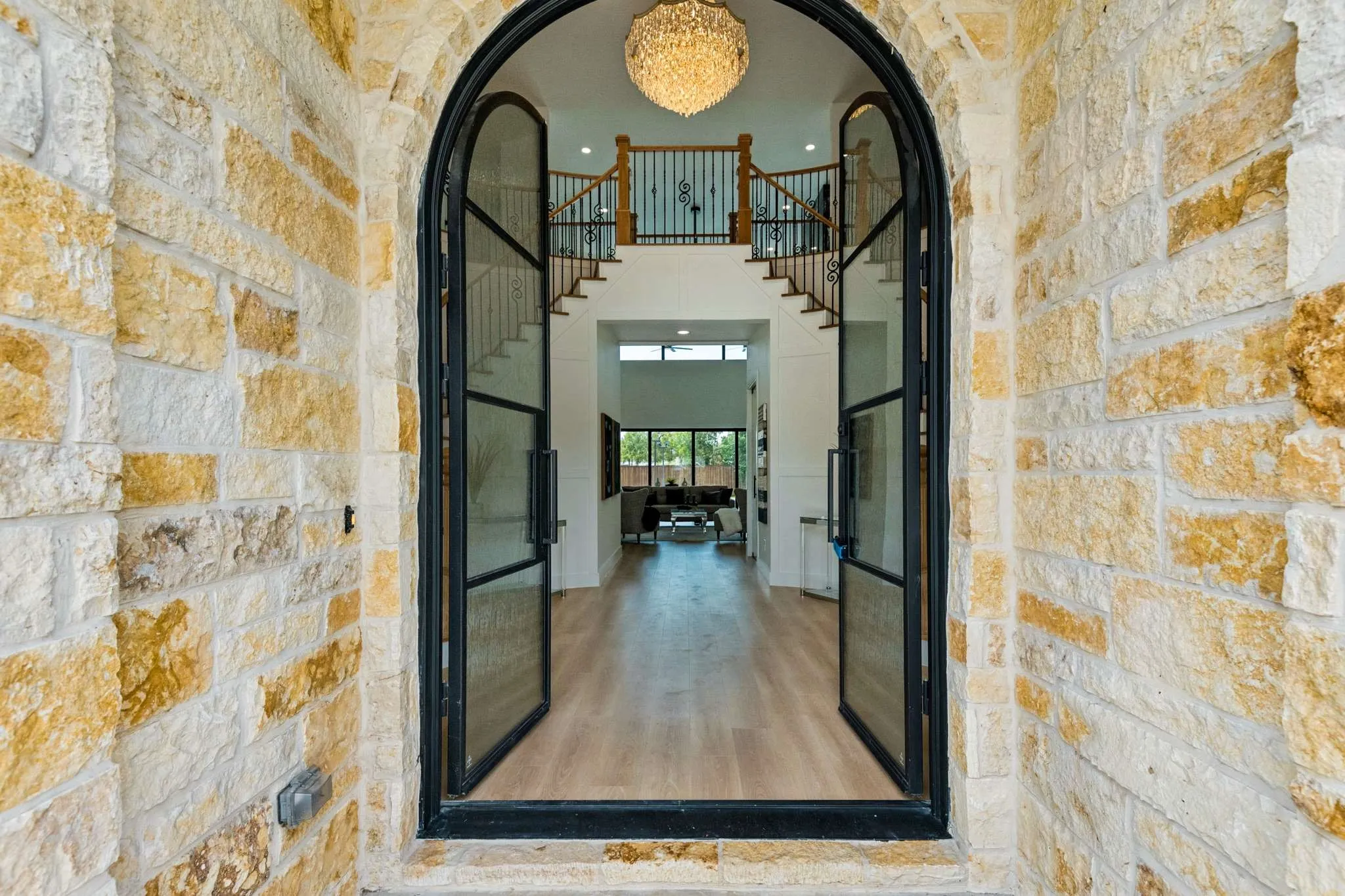 Arched stone entryway featuring double glass doors, a prominent chandelier, wood-finish flooring, and a grand staircase with wrought iron railing