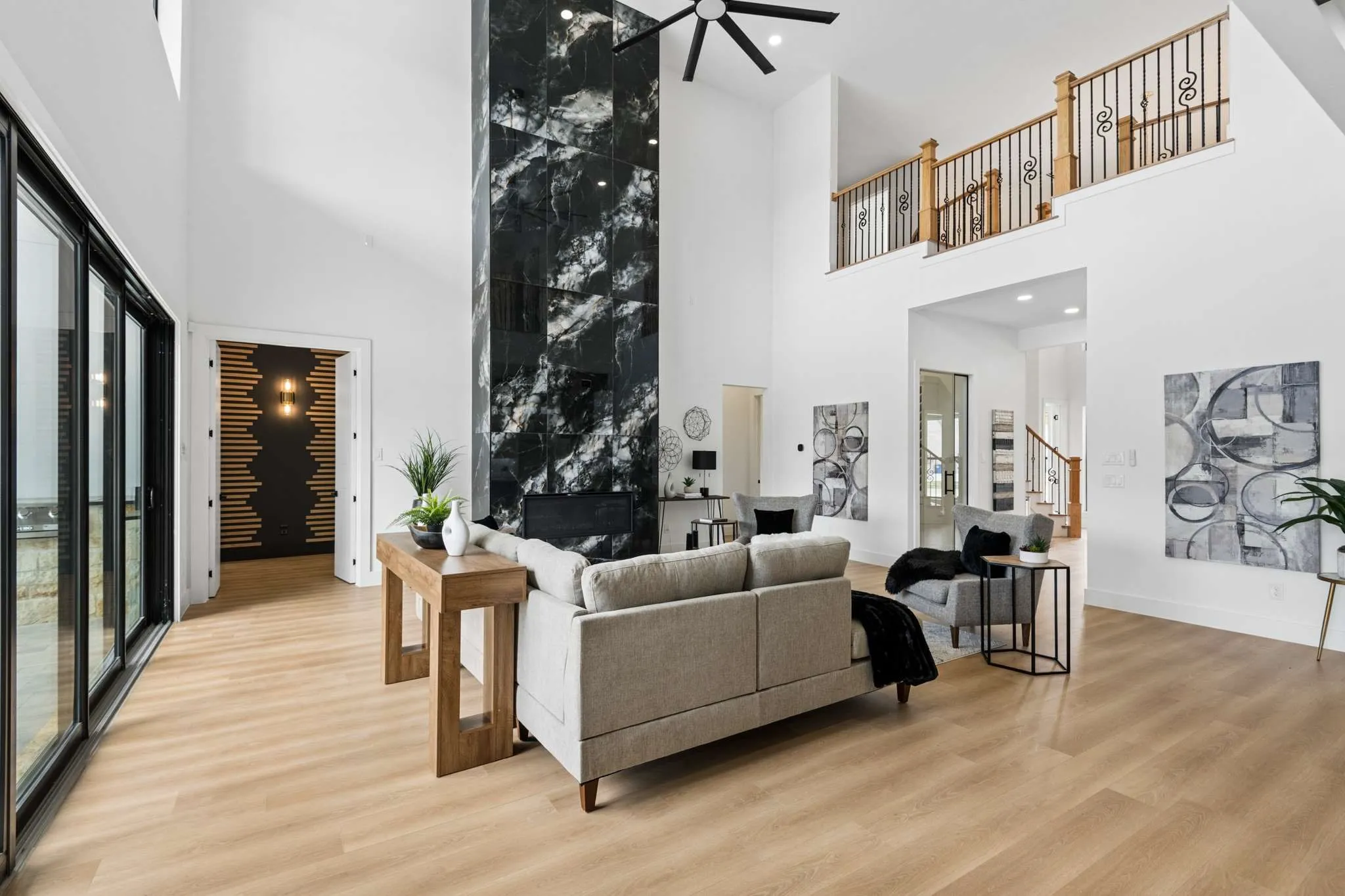 Spacious living area featuring high vaulted ceilings, expansive sliding glass doors, a prominent black stone fireplace, wood-finish flooring, and a decorative upper-level railing