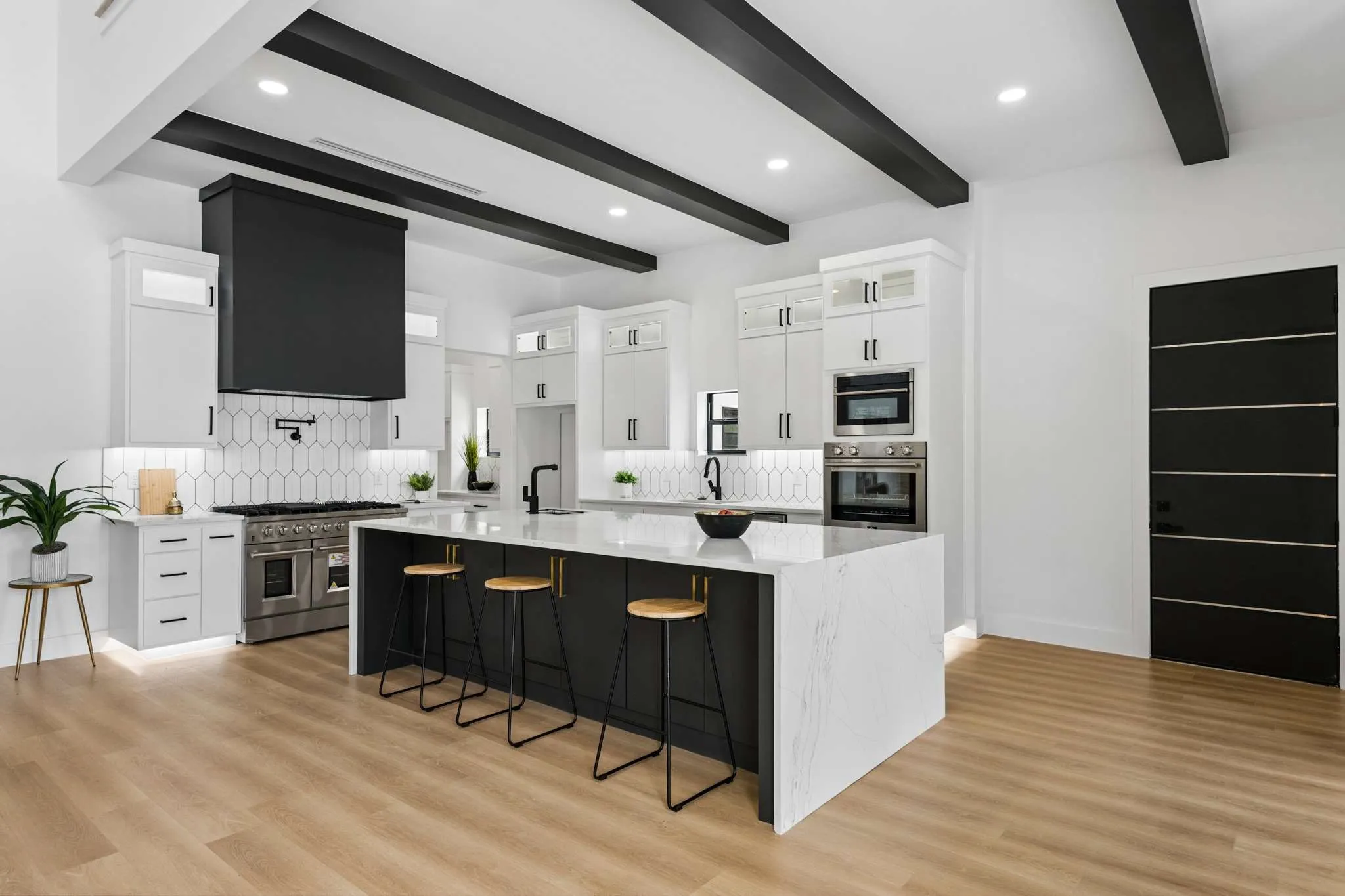 High ceilings with exposed dark beams, wood-finish flooring, white cabinetry with dark hardware, a large kitchen island with a light-toned countertop, and stainless steel appliances