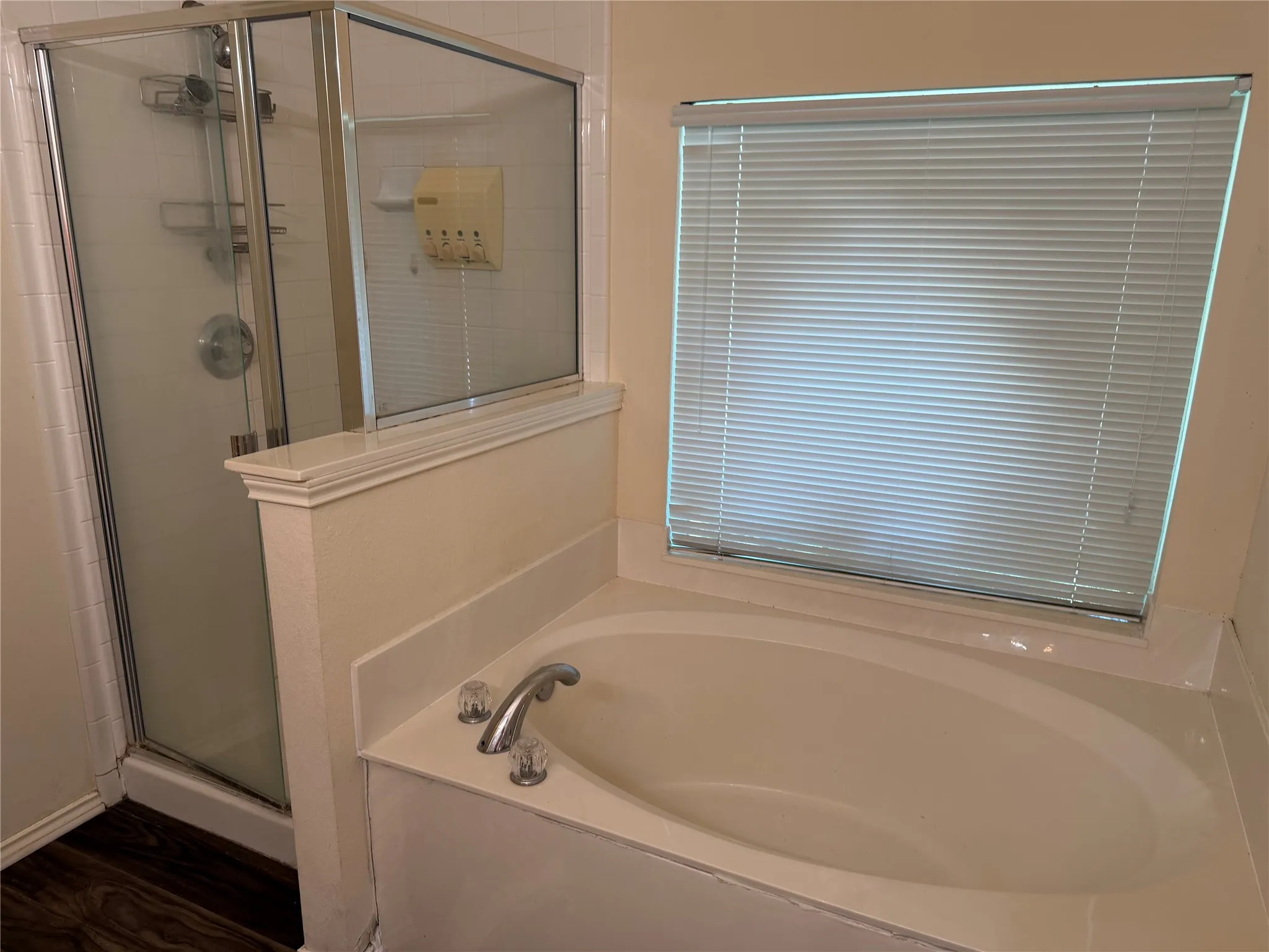 Built-in soaking tub with chrome fixtures, glass-enclosed shower with a tiled surround, and a privacy window with blinds