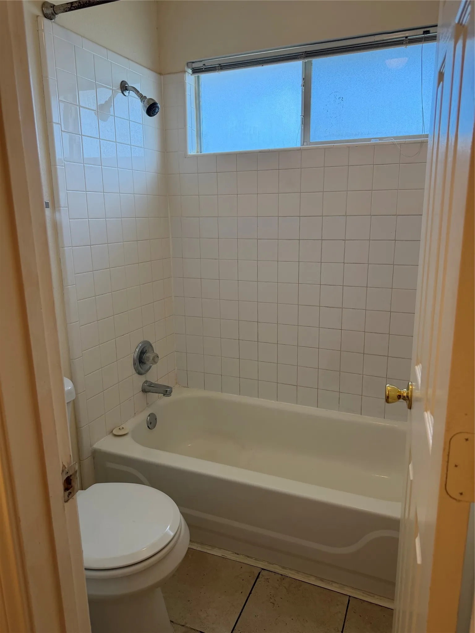Bathtub with tile surround, frosted glass window, wall-mounted showerhead, and tile flooring