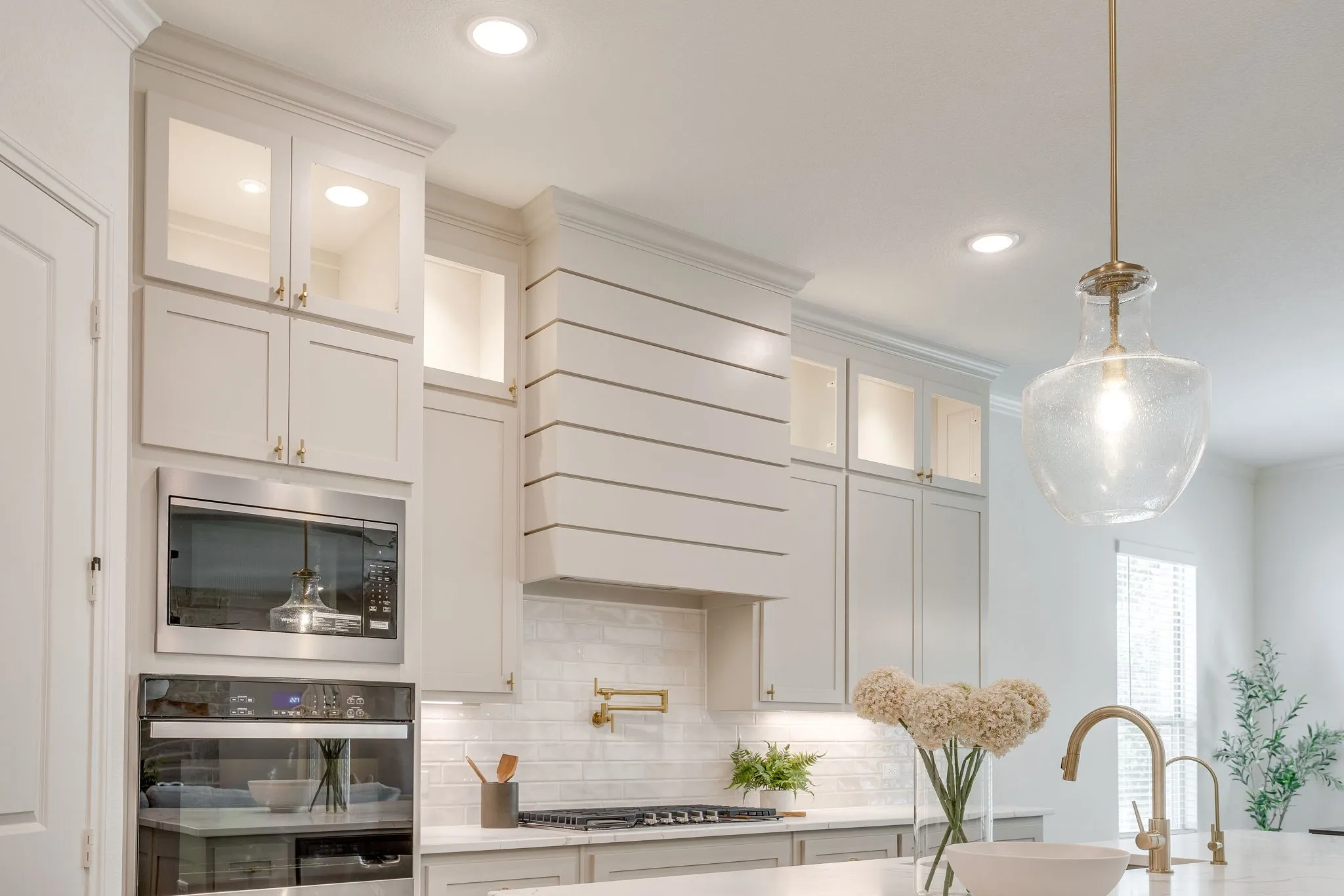 White shaker-style cabinetry with glass-front uppers, stainless steel built-in microwave and oven, shiplap-accented range hood, and a clear glass pendant light