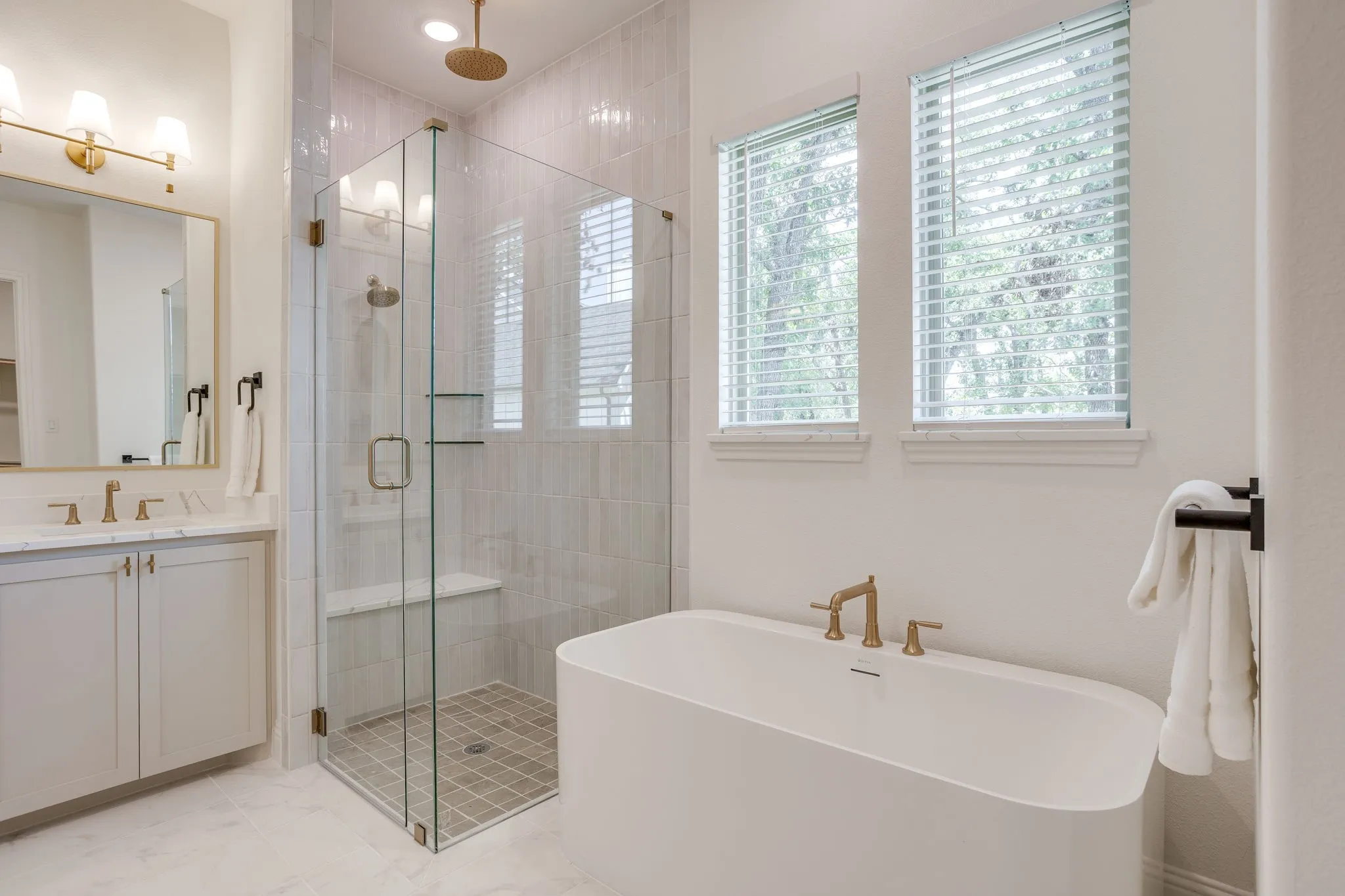Freestanding soaking tub with brass fixtures, glass-enclosed shower with bench and built-in shelving, dual-sink vanity with brass fixtures and stone countertop, and two windows with blinds
