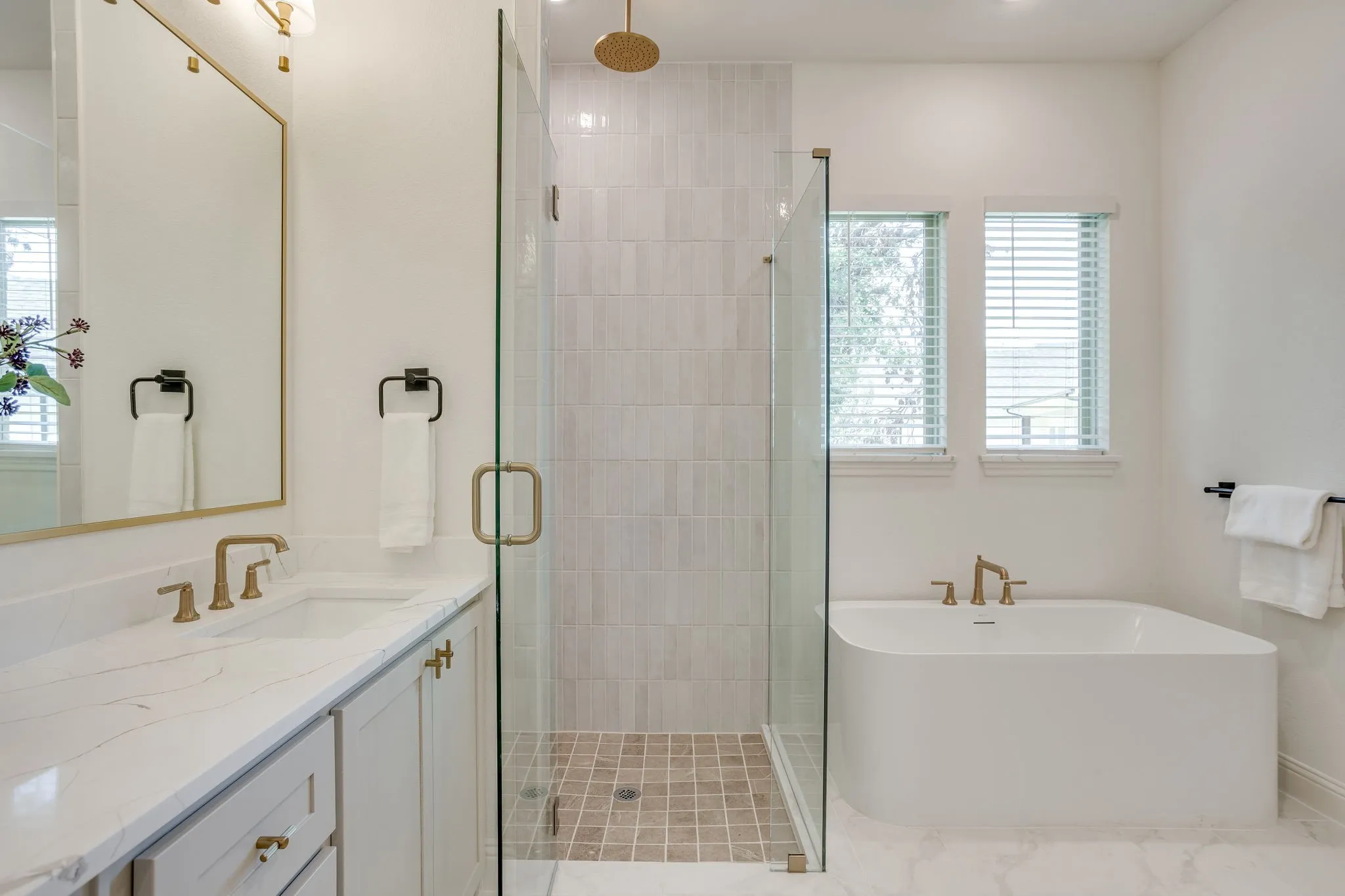 Freestanding soaking tub, glass-enclosed shower, dual windows, vanity with integrated sink, and gold-toned fixtures