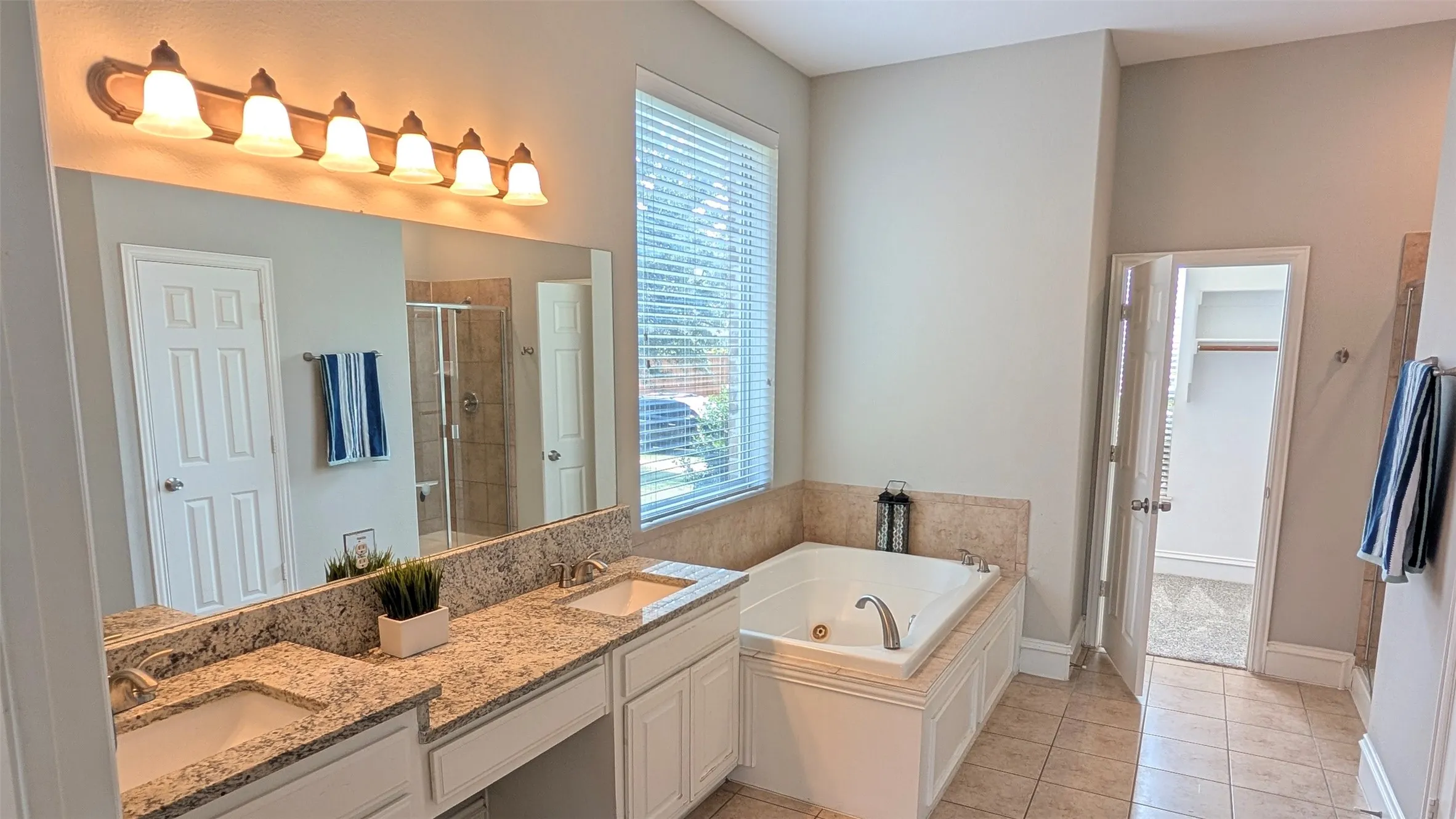 Dual sink vanity with granite-style countertop, large mirror, built-in bathtub, and tiled flooring