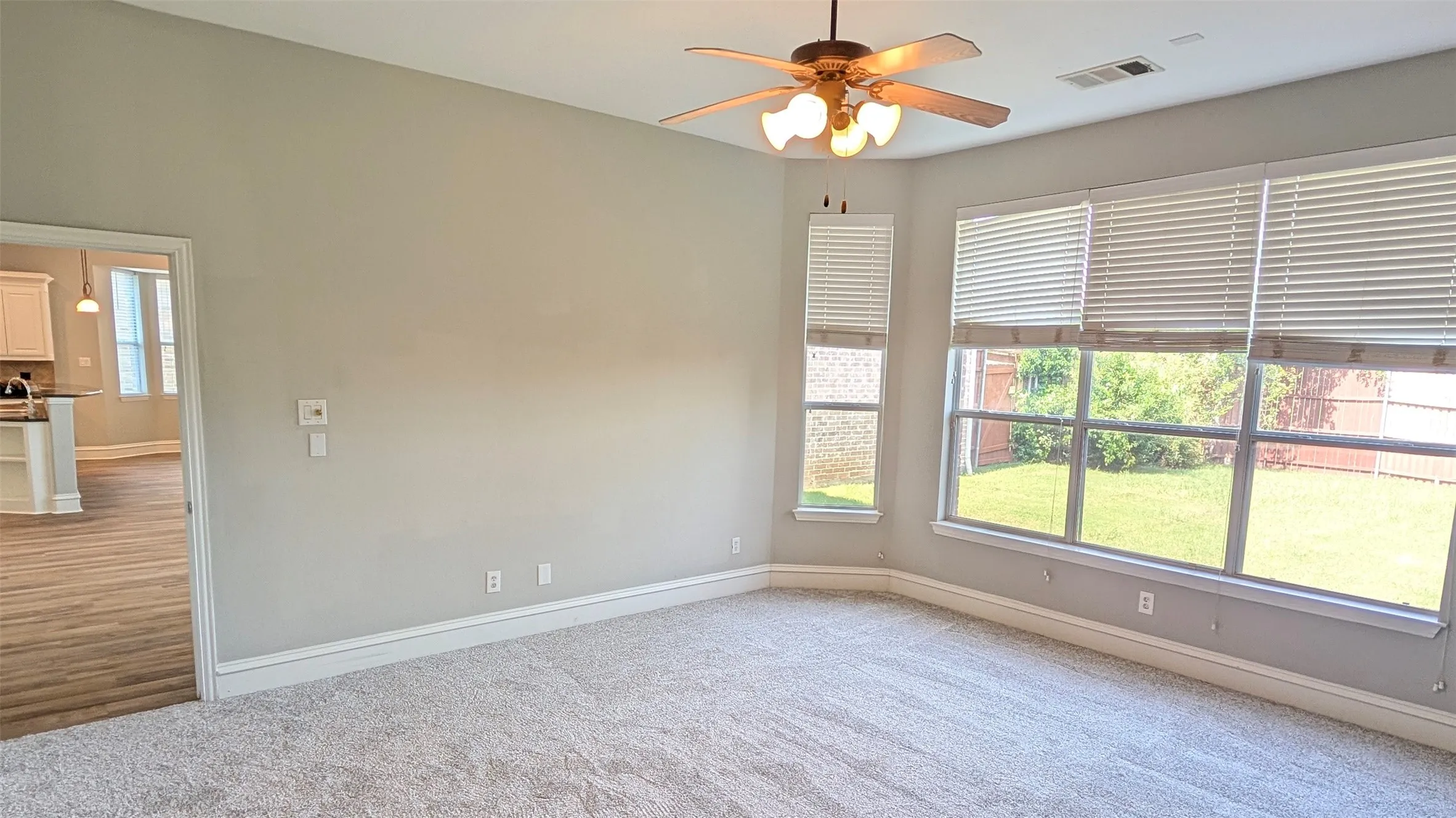 Spacious room featuring light-toned carpeting, multiple windows with blinds, a ceiling fan with lighting, and a doorway opening to wood-finish flooring