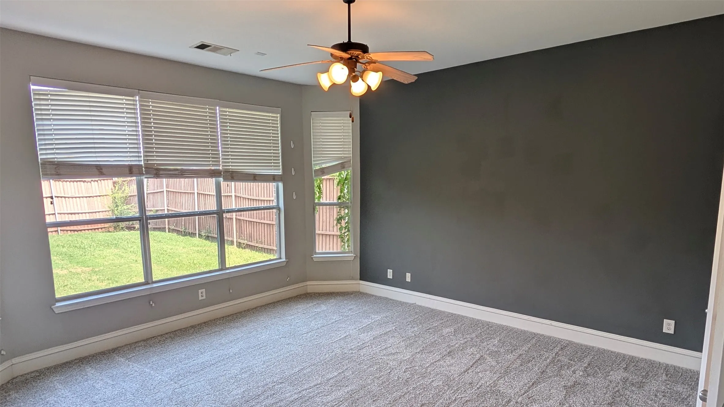 Corner windows with integrated blinds, ceiling fan with light fixture, gray accent wall, light gray wall paint, and gray carpet flooring