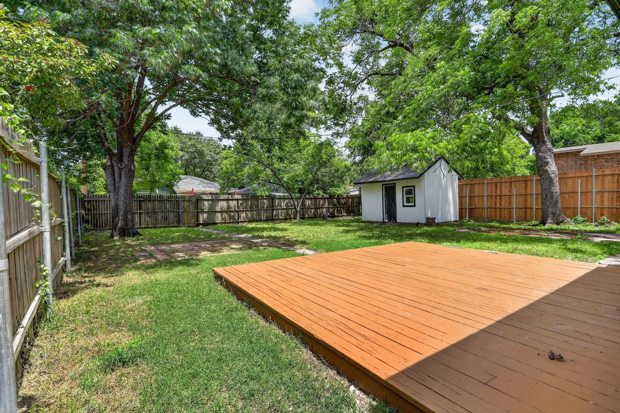 Expansive backyard featuring a large wood deck, a white storage shed with a dark door, and mature shade trees