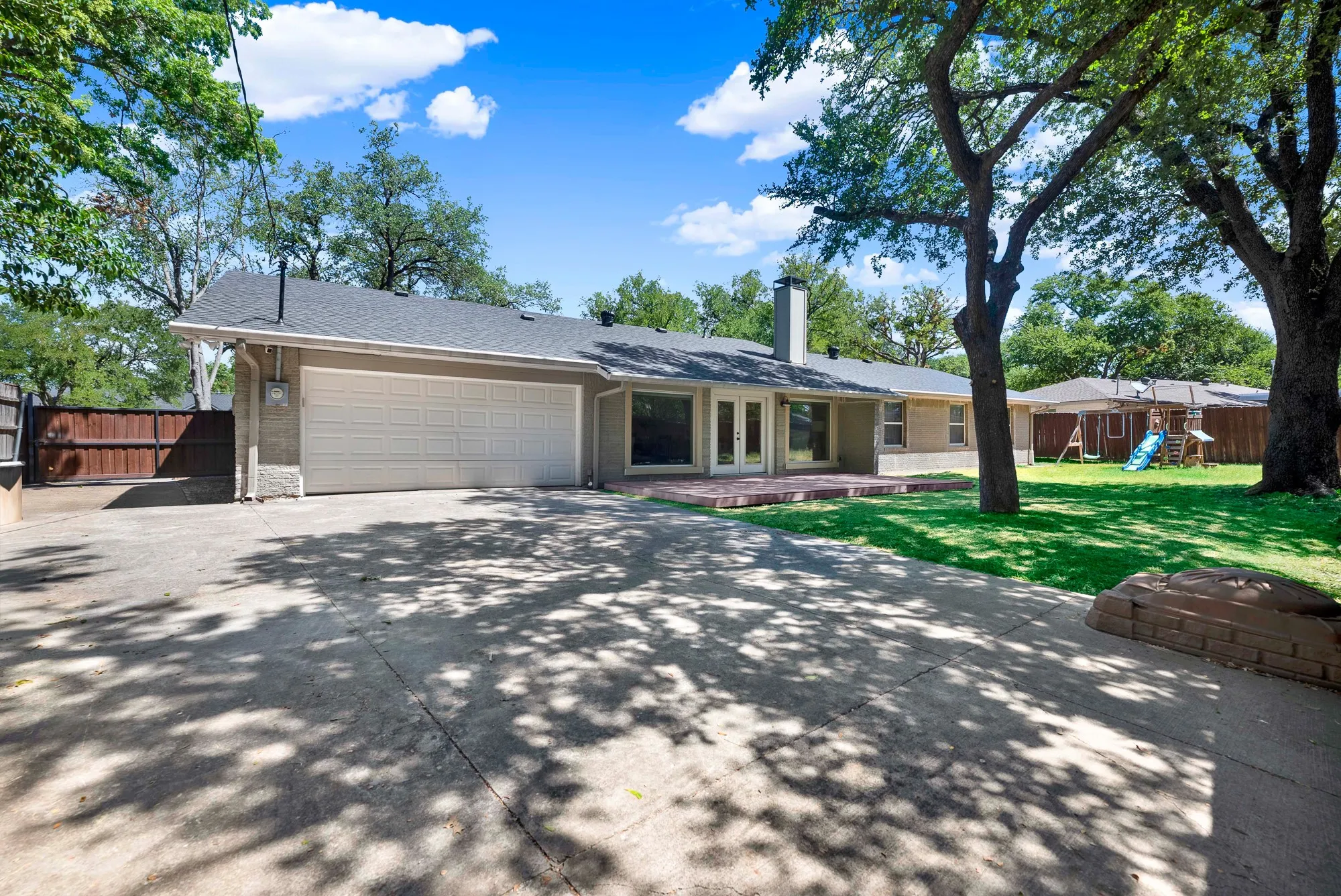 Attached two-car garage, expansive concrete driveway, mature trees providing shade, brick patio, and a large grassy backyard