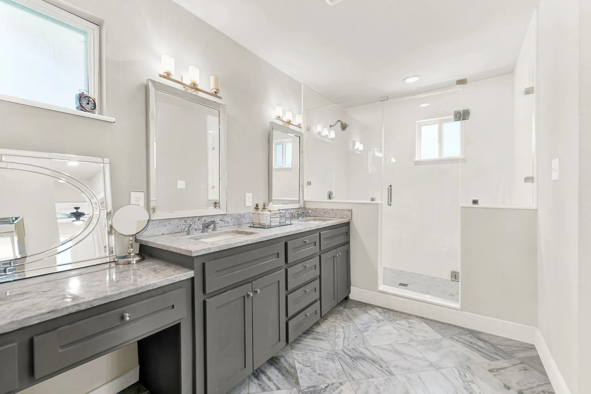 Dual vanity with gray cabinetry and light-toned countertops, walk-in shower with glass enclosure, large format tile flooring, and multiple windows providing natural light