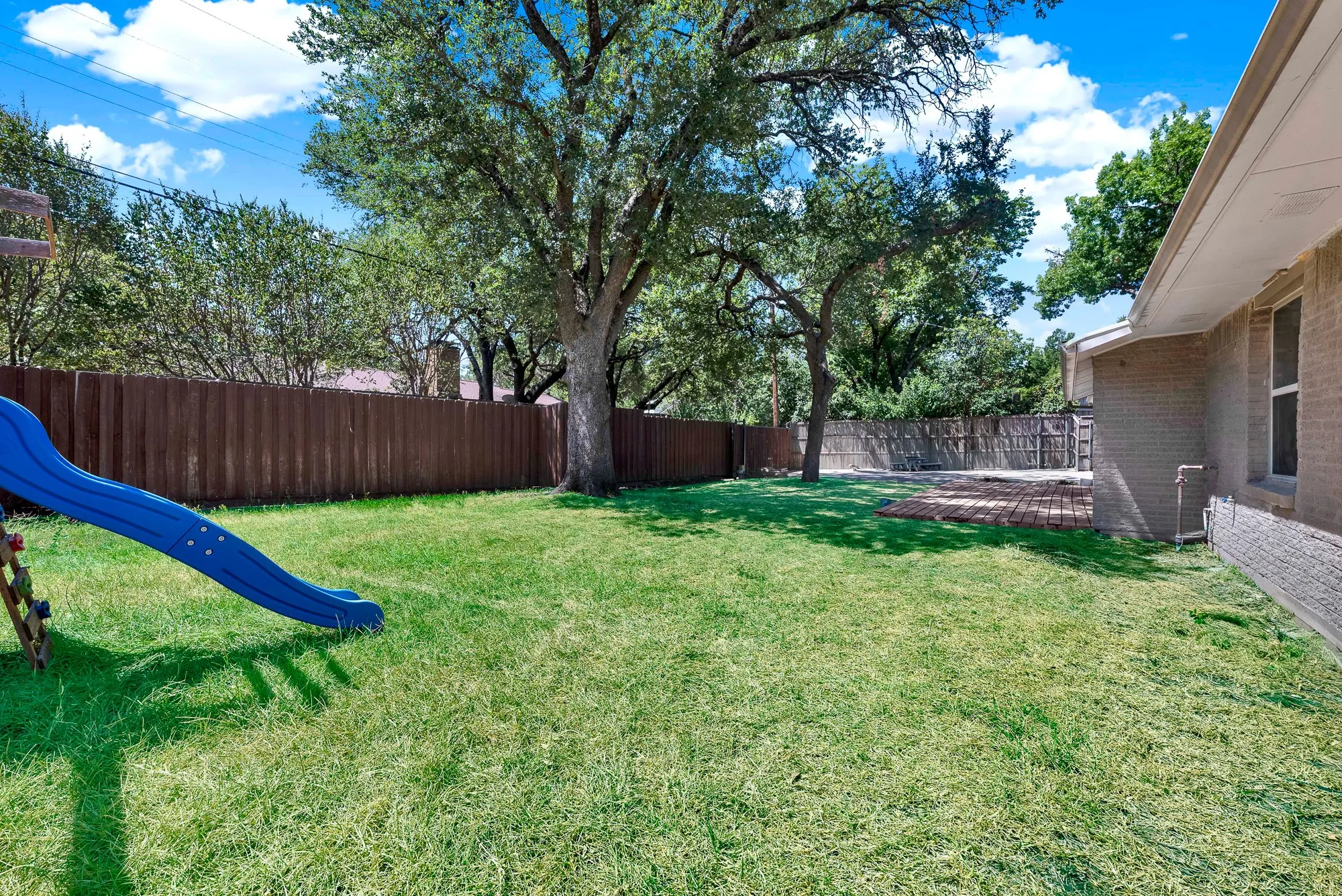 Expansive grassy yard with mature trees, a privacy fence, and brick paver patio