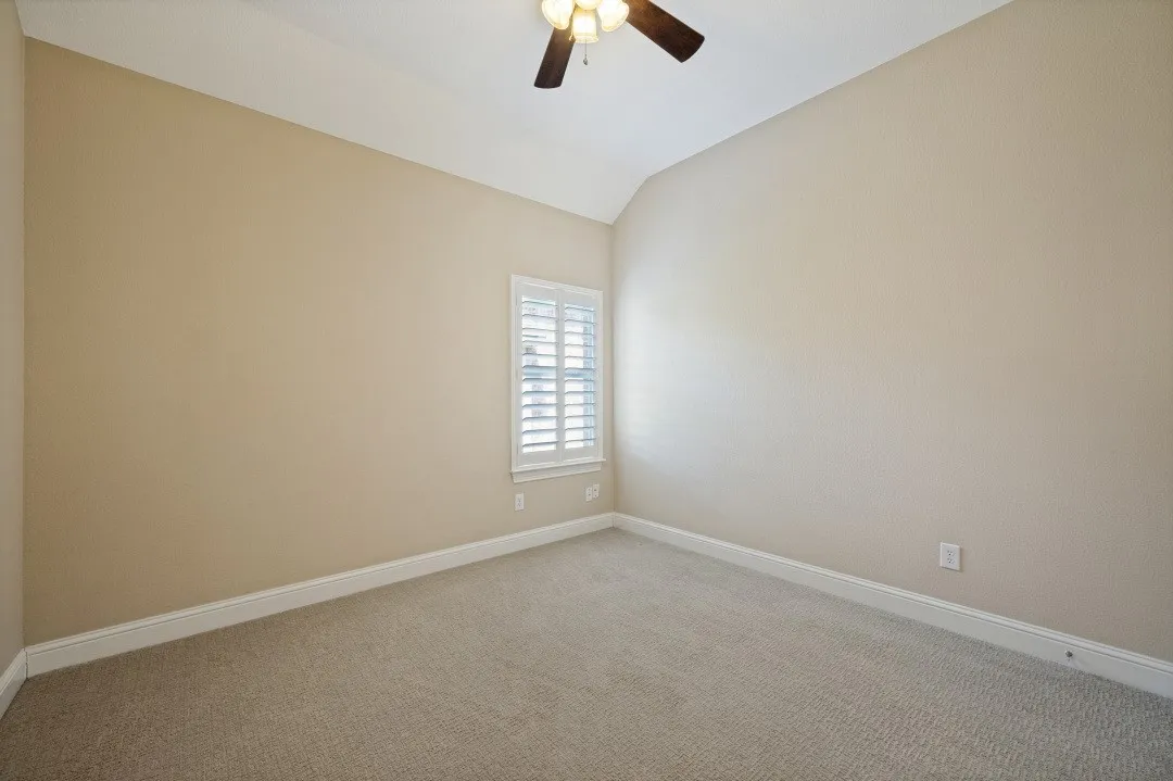 Vaulted ceiling, ceiling fan, neutral carpet, single window with white plantation shutters, and light-toned walls
