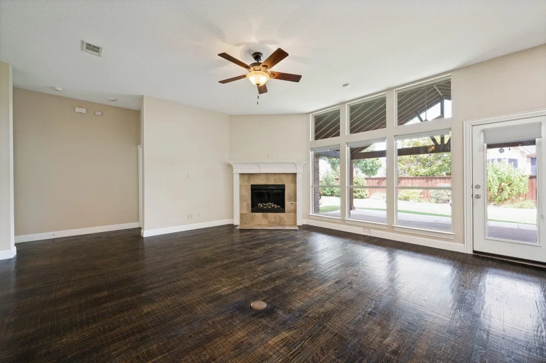 Expansive living area featuring large picture windows, dark wood-finish flooring, a corner fireplace with a tiled surround, and a ceiling fan