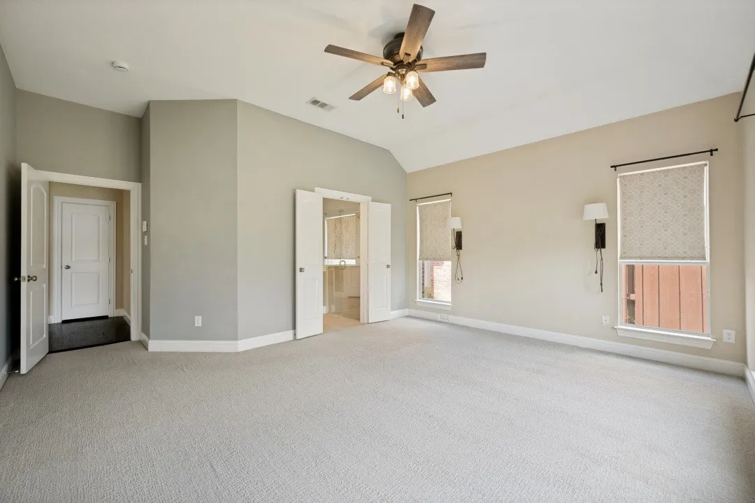 Vaulted ceiling, ceiling fan, two windows with roller shades, light-colored carpeting, and painted walls