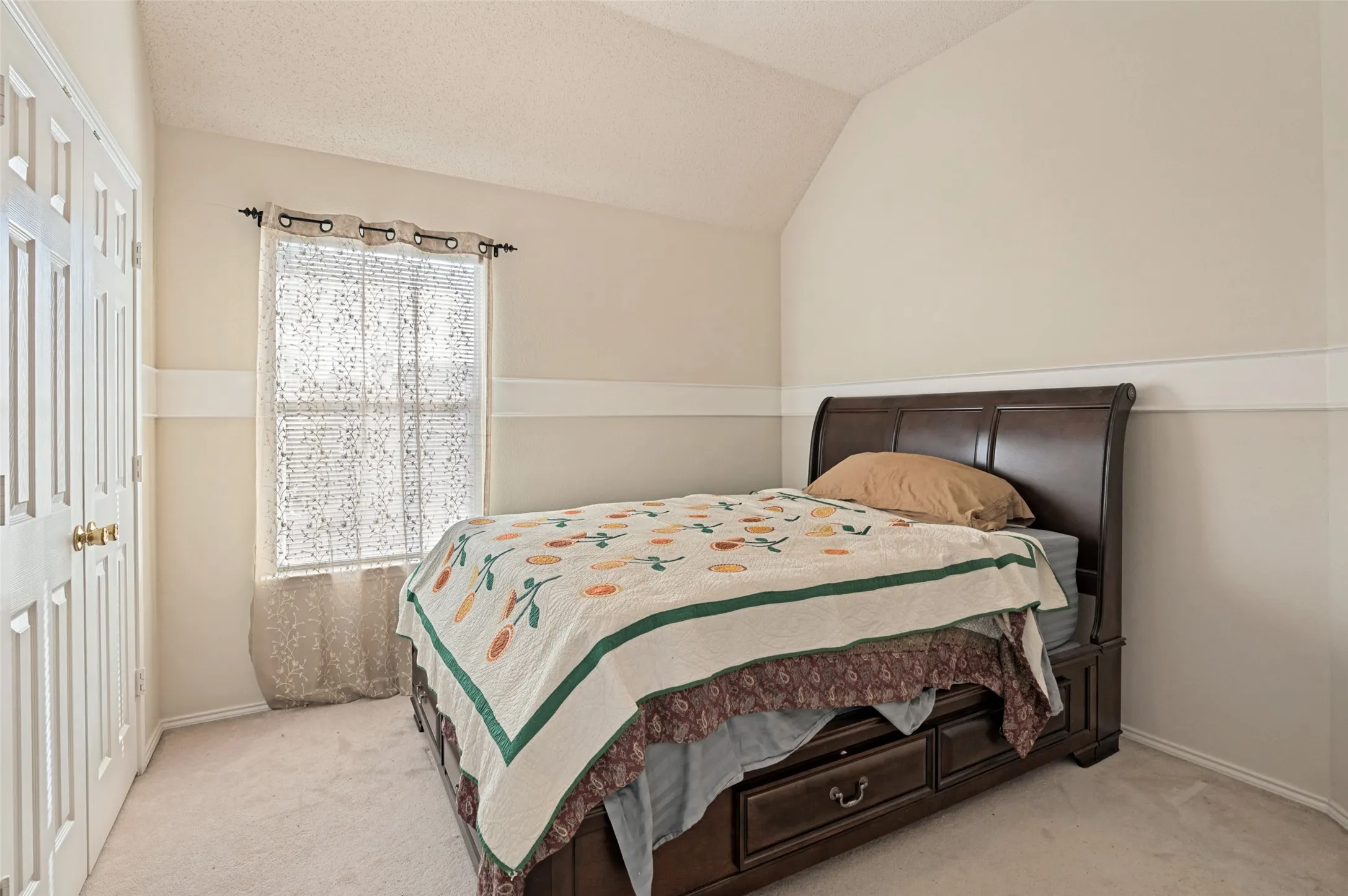 Vaulted ceiling with textured finish, light-colored carpeting, white paneled bifold closet doors, and a window with a valance